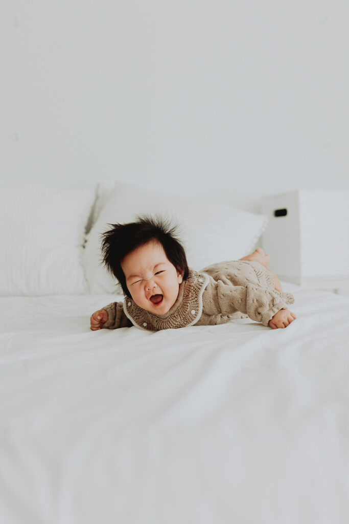 photo of a 3 month old baby laying on her stomach on a big bed with white sheets. she has a huge smile at the camera. taken in langley