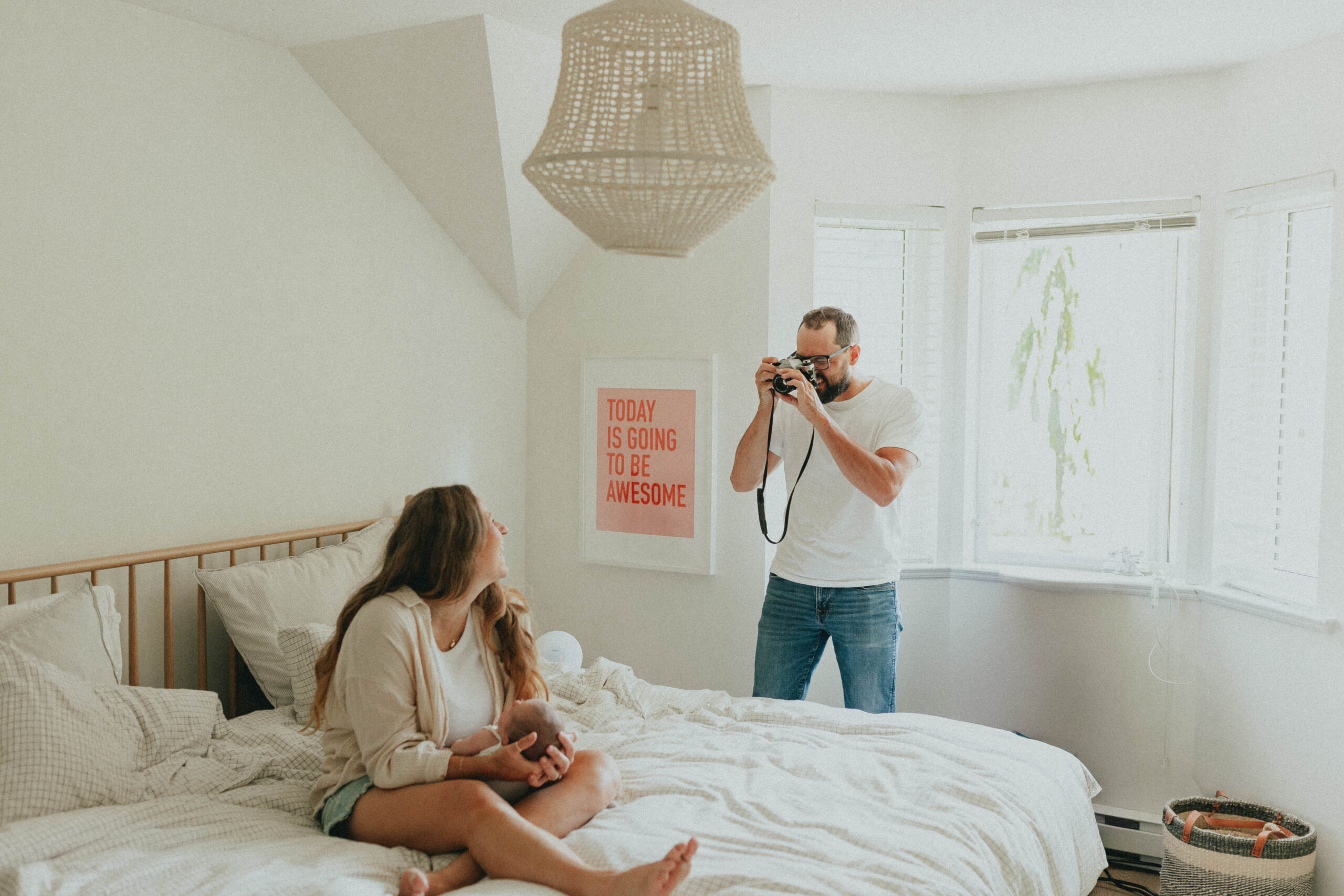 photo of a mom sitting on a bed with dad standing beside the bed taking a picture with a film camera while mom holds their newborn baby taken in vancouver