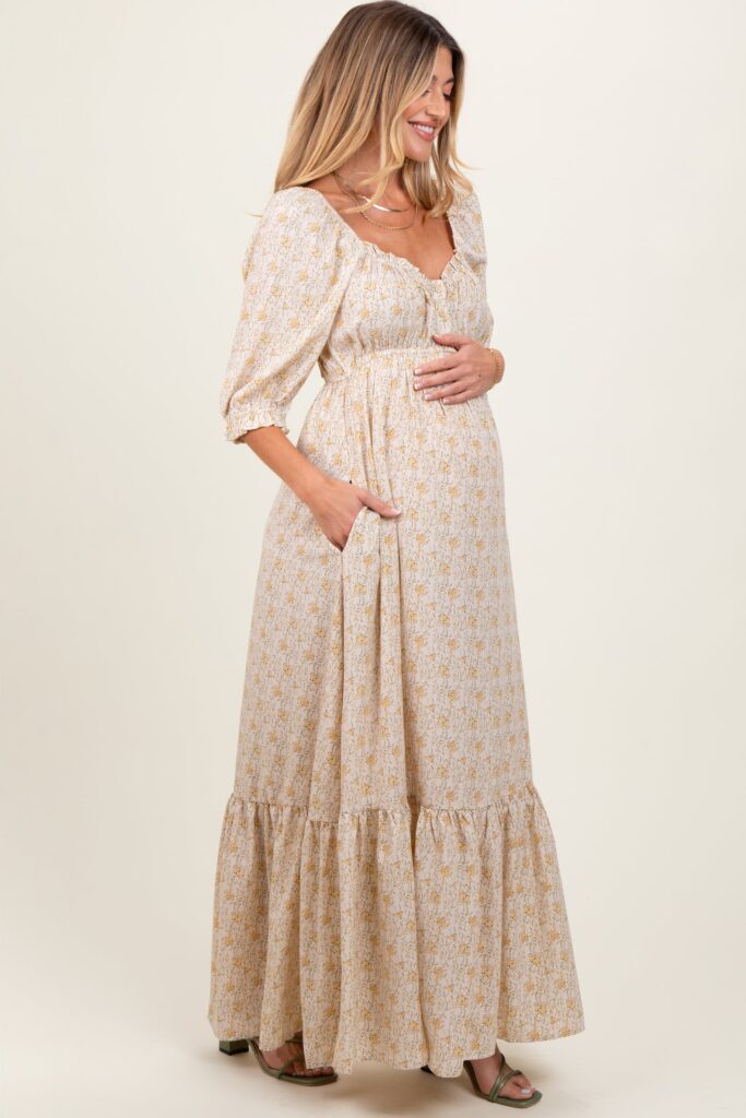 Photo of a model cradling her baby bump while wearing a cream and floral patterned quarter sleeve maternity photoshoot dress taken on a white background