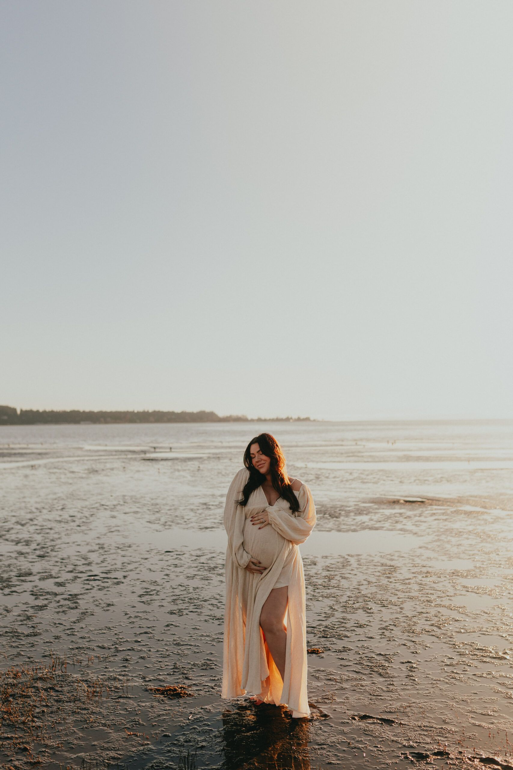 photo of a pregnant woman standing in the low ocean water cradling her bump while looking down with her eyes closed