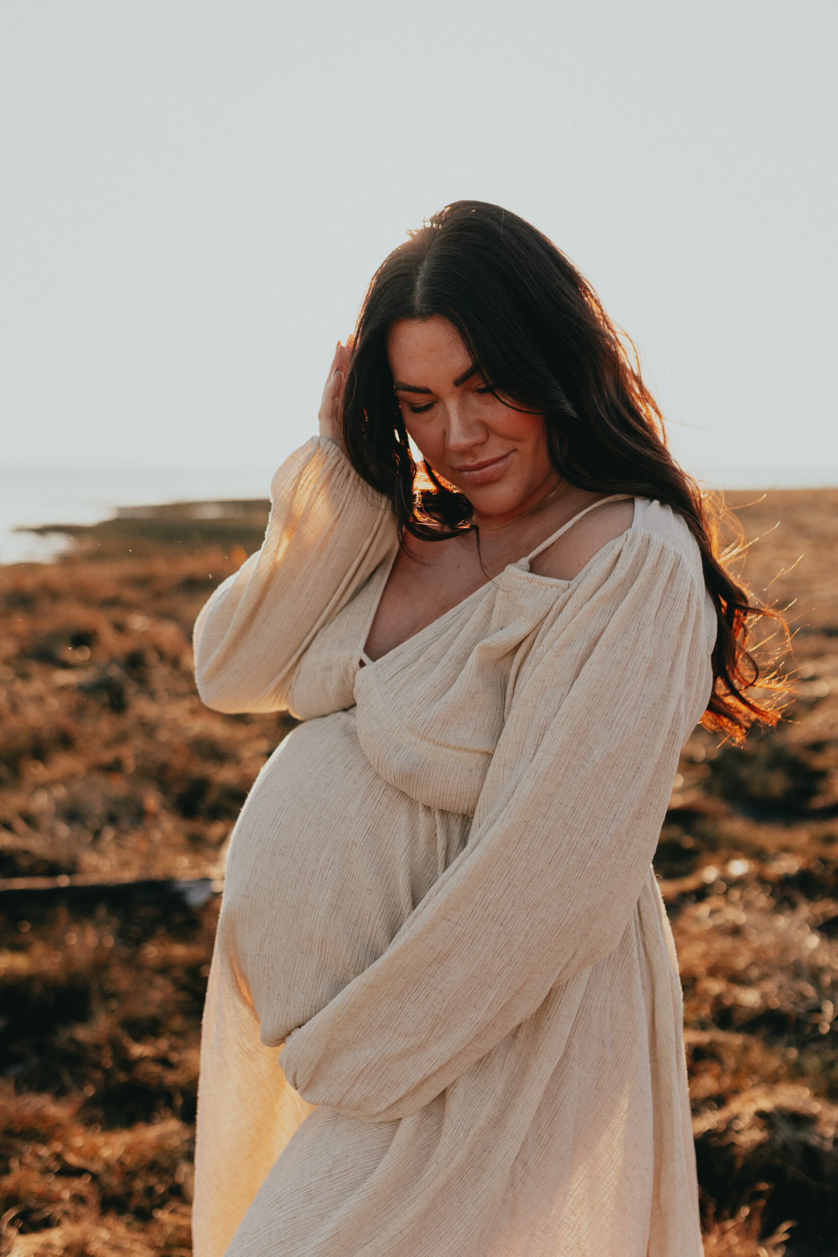 photo of a pregnant woman on the beach holding her hair with one hand and her bump with the other while looking down with her eyes closed