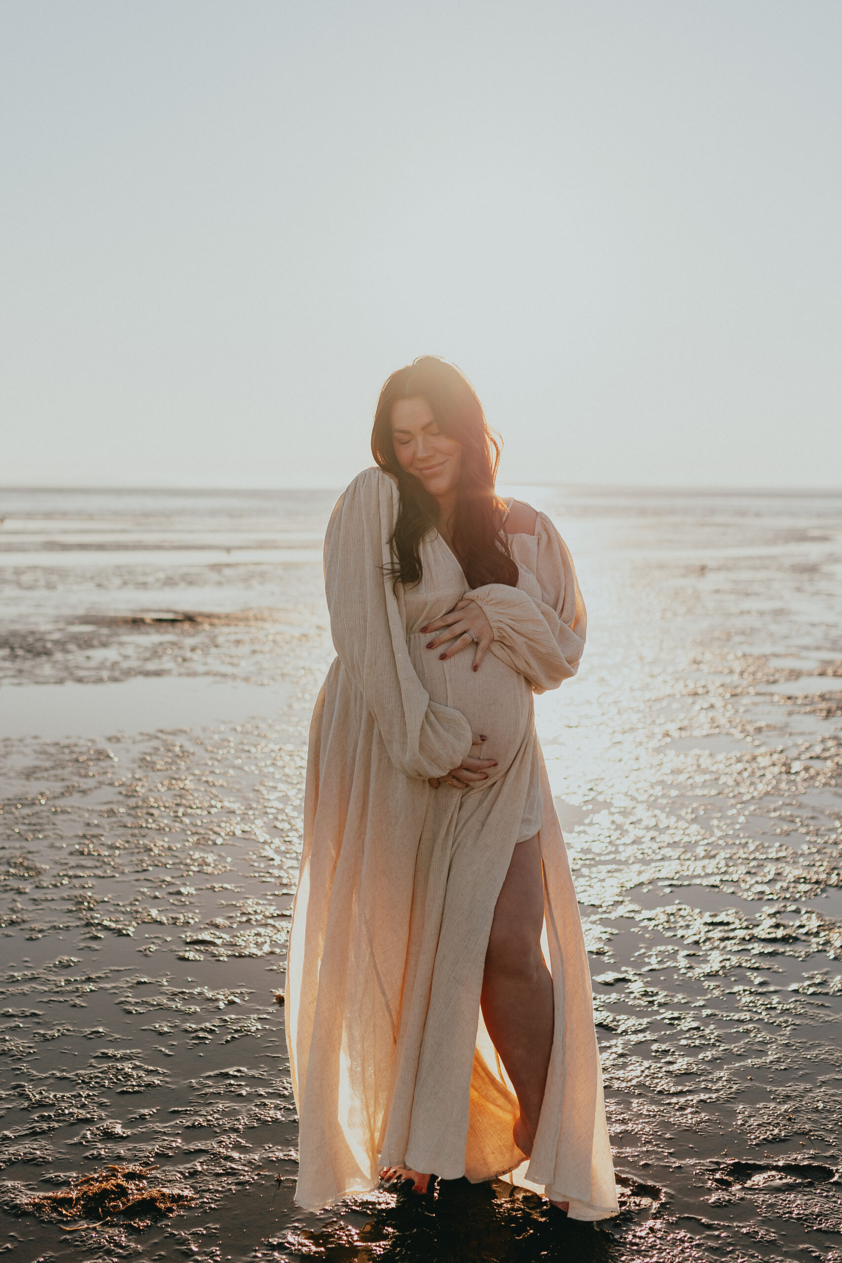 photo of a pregnant woman standing on the beach cradling her bump while looking down with her eyes closed