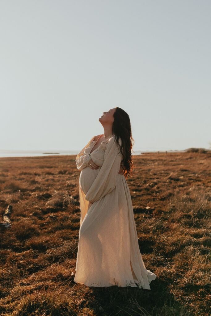 photo of a pregnant woman holding her belling while holding her head back with her eyes closed taken on the beach