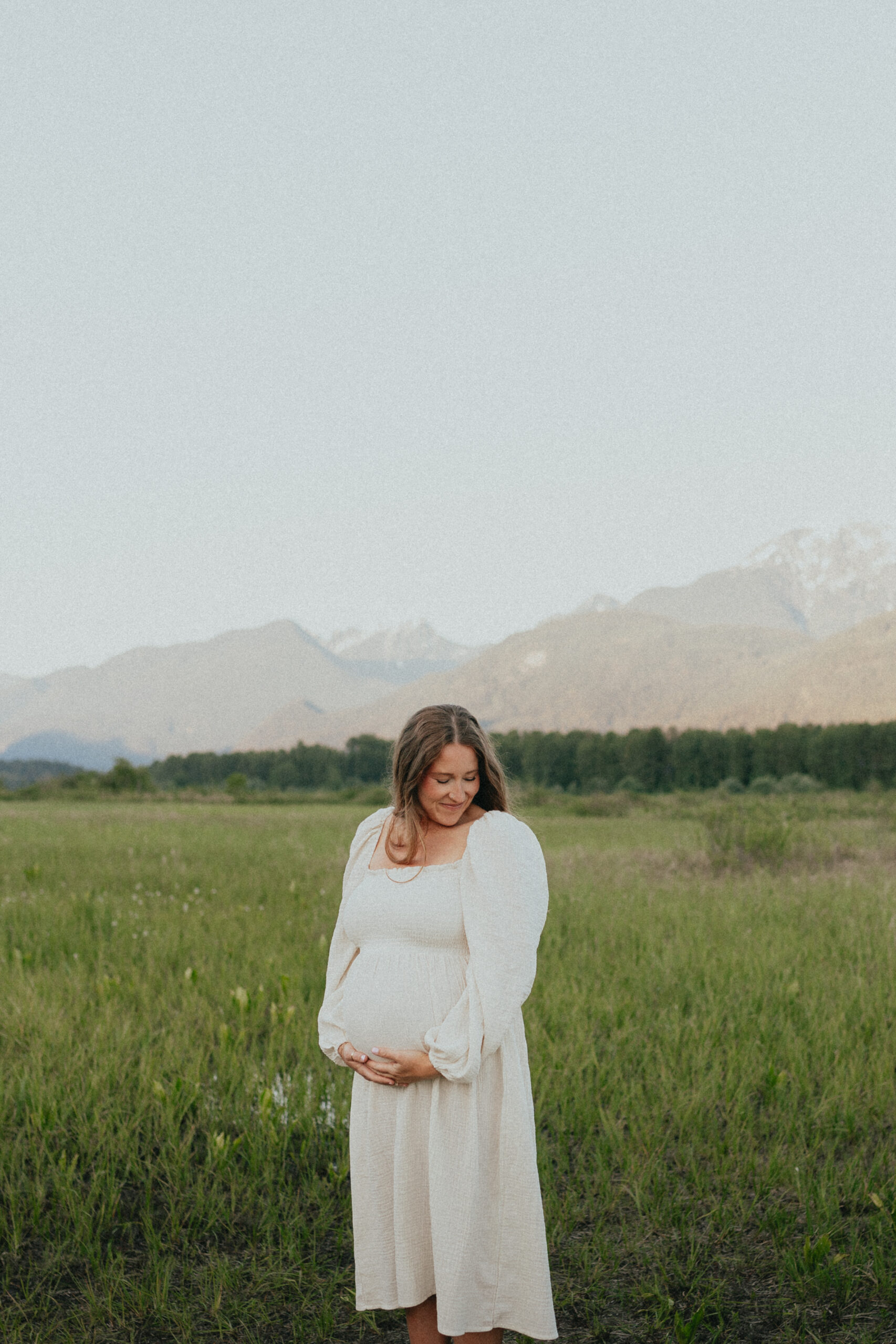 photo of a pregnant woman standing in a field with mountains behind her while she cradles her belly and looks down smiling