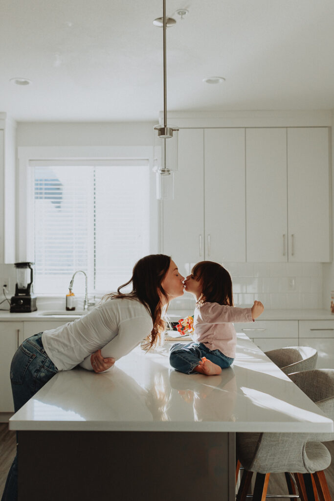 family photography photo of a mom leaning in to kiss her daughter who is sitting on the kitchen island while holding her arms back taken in langley