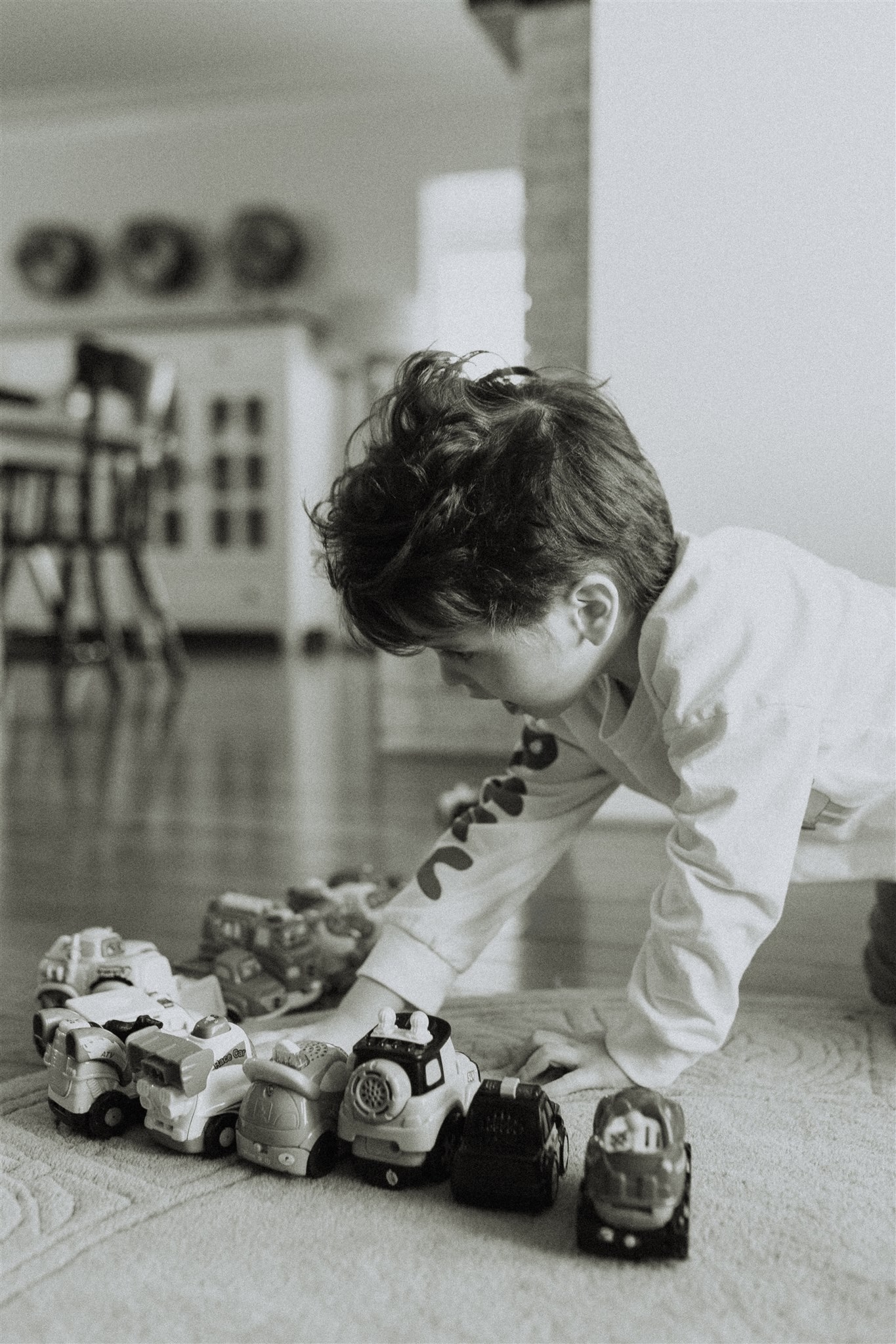 Black and white photo of a little boy reach for his toy cars that are all lined up along the ground. 