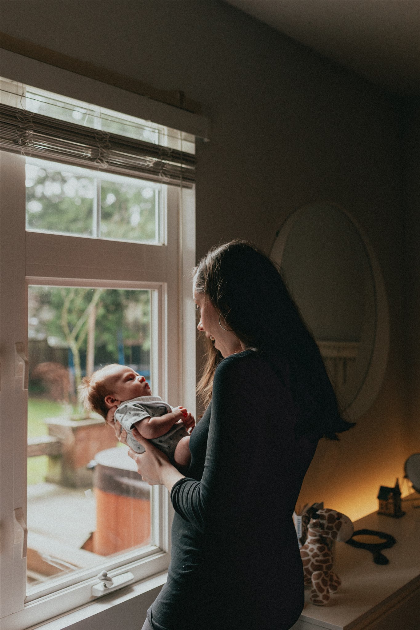 photo of a mom holding her newborn baby taken from behind but you can see mom smiling at her baby while looking at him