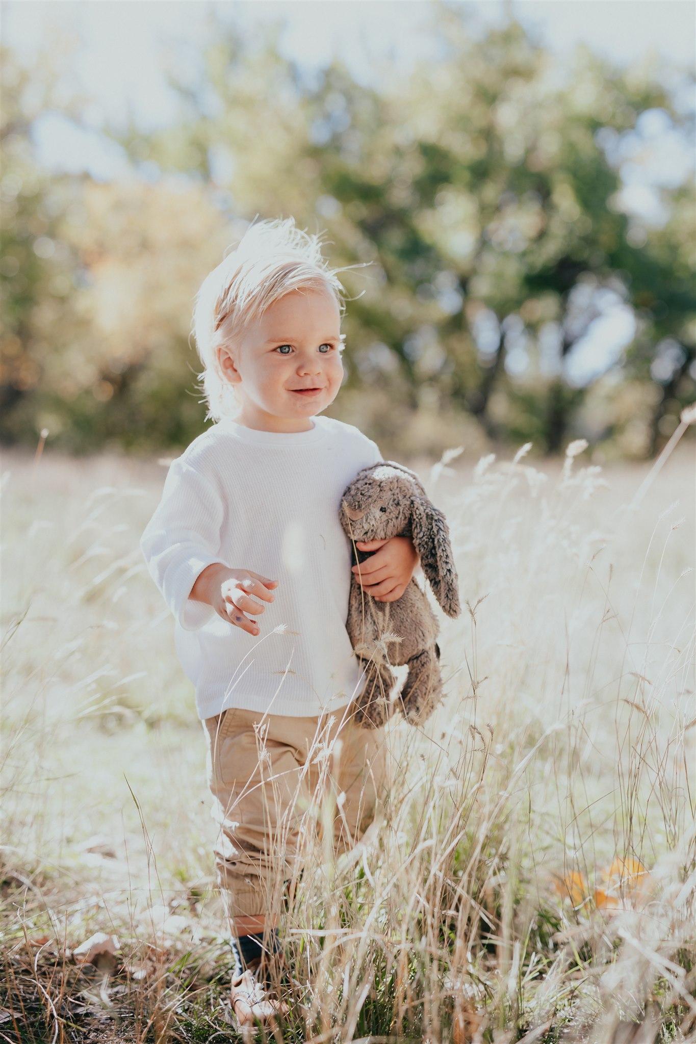 photo of a little boy standing in a field holding a stuffed bunny smiling and his parents off camera