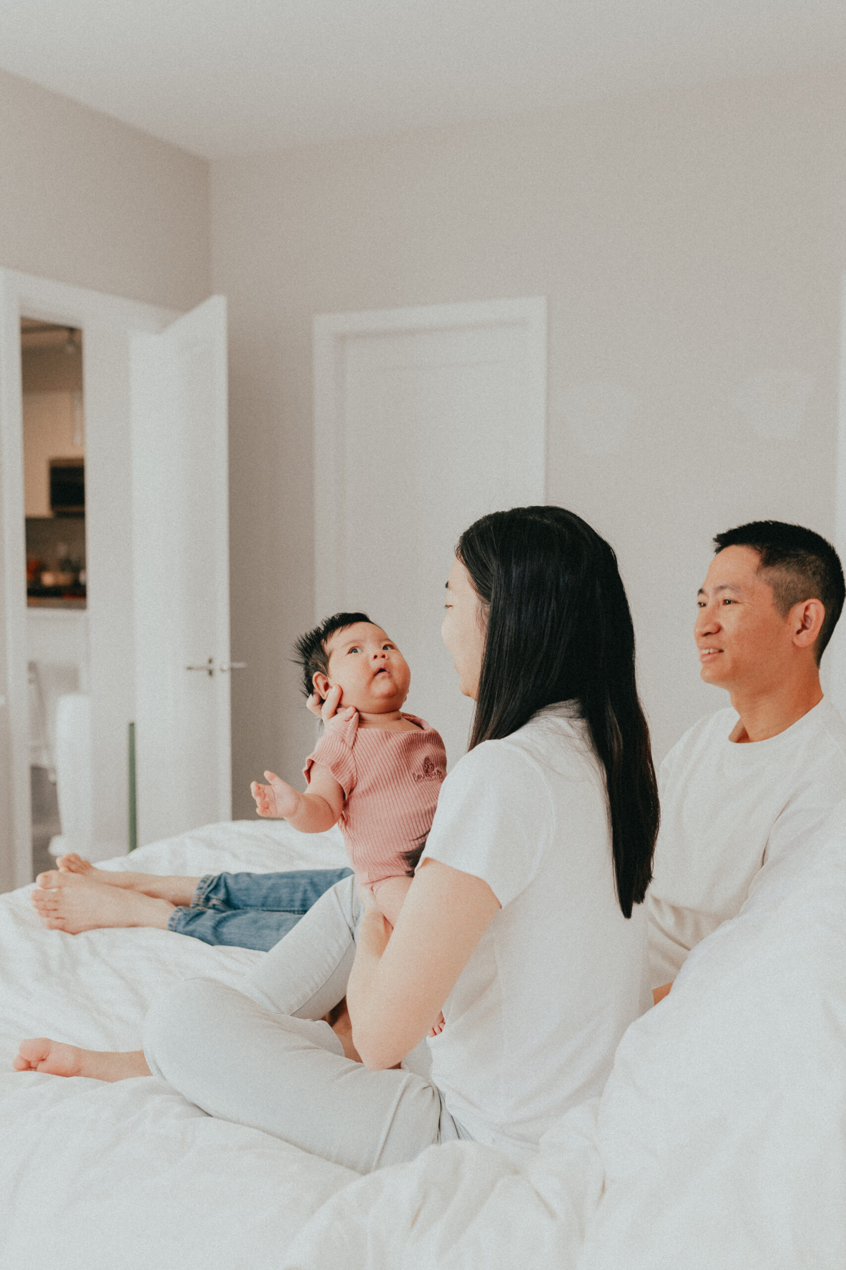 photo of a mom and dad sitting on a bed with mom holding her newborn baby and smiling at her. taken by a langley newborn photographer