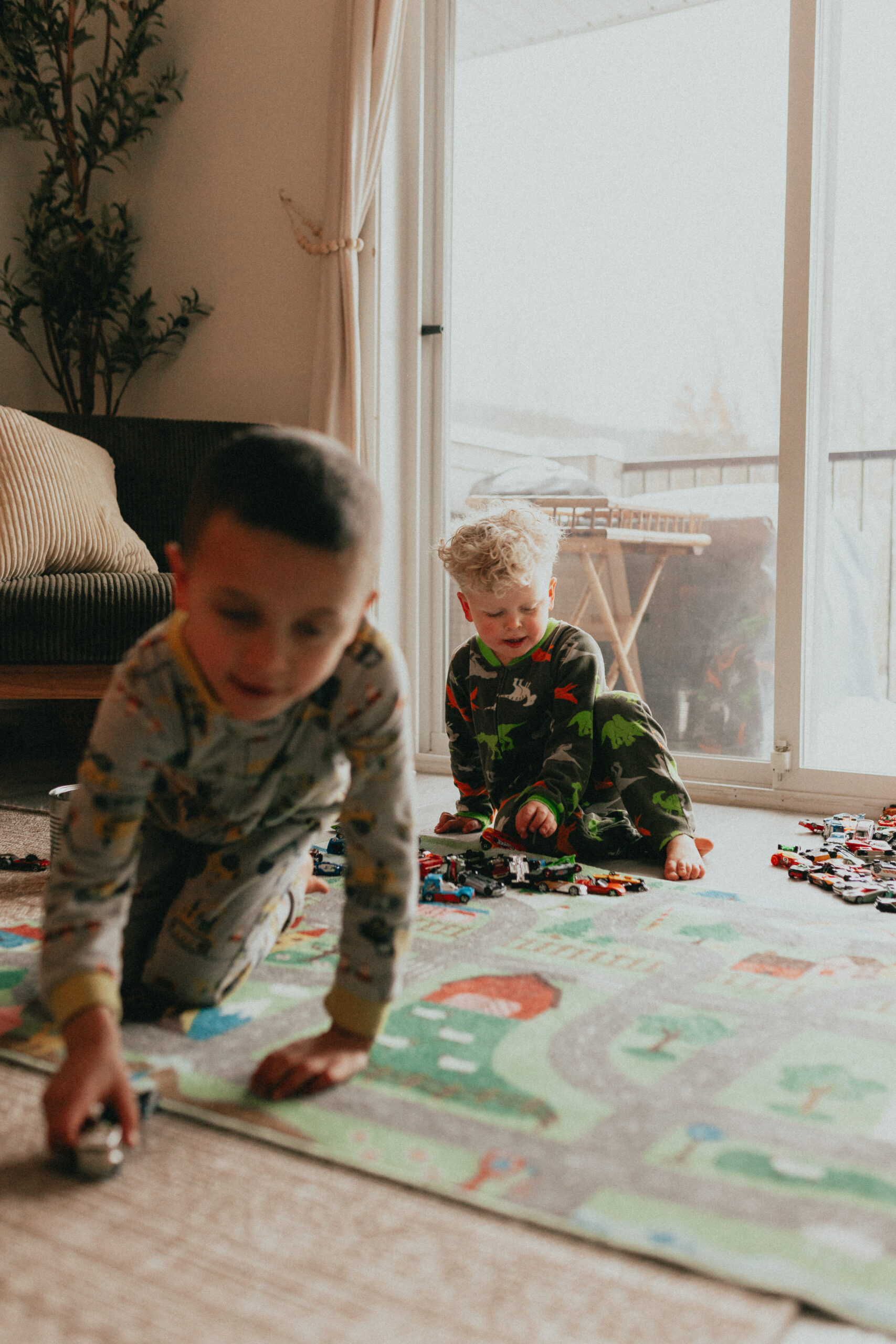 Photo of 2 little boys playing cars with the boy sitting farther away in focus looking down at his cars