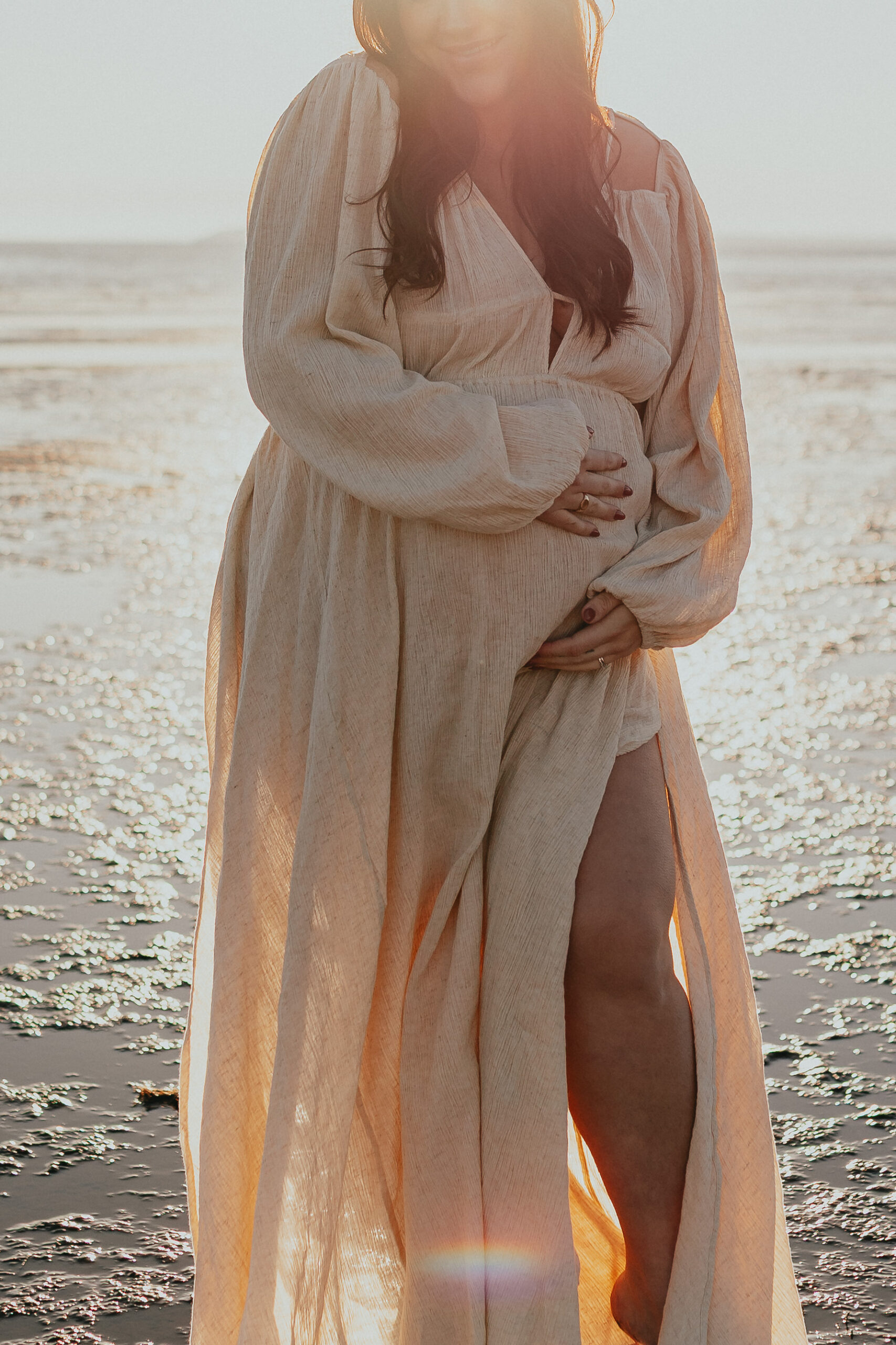 Photo of a pregnant woman on the beach with the sun behind her cradling her baby bump and smiling taken by a vancouver maternity photographer