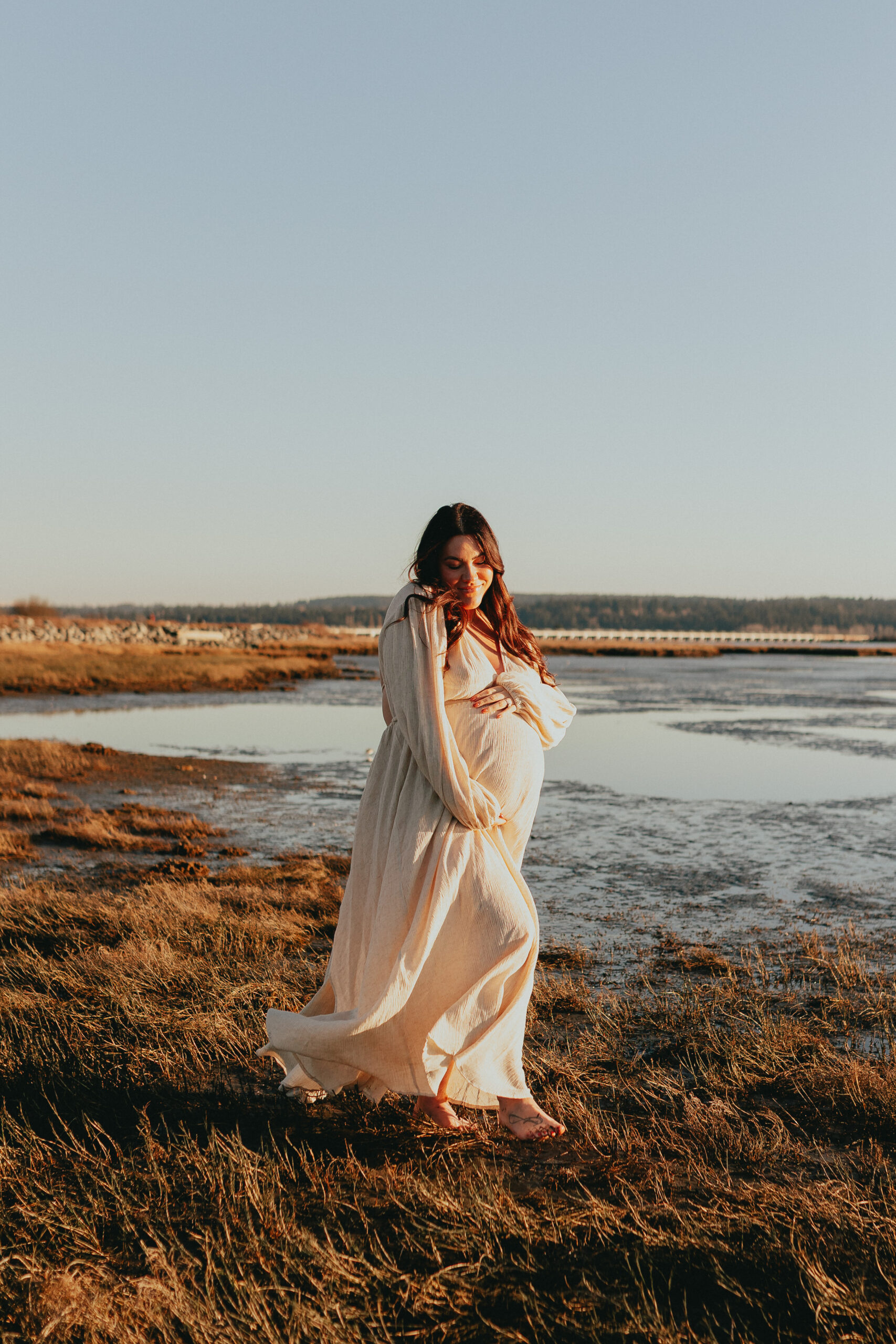 Photo of a pregnant woman on the beach wearing a beige flowing dress and holding her baby bump while walking and looking down and smiling