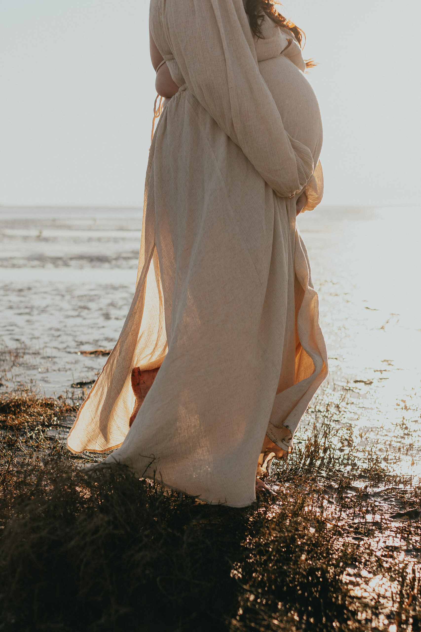 Photo of a pregnant woman wearing a beige flowing dress walking along the beach where you can only see from her shoulders down taken by a vancouver maternity photographer