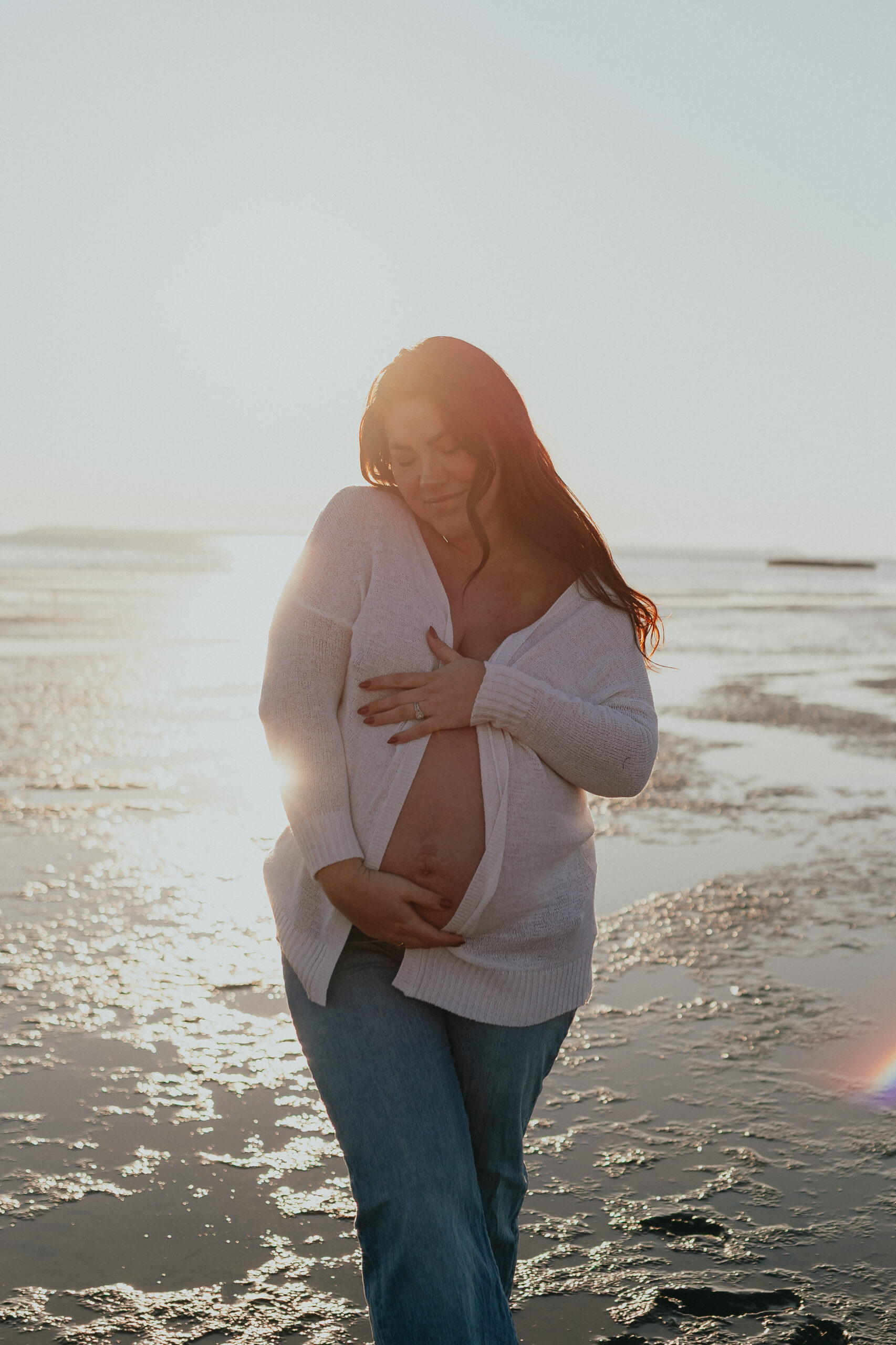 Photo of a pregnant woman holding her baby bump and looking down and smiling while standing on the beach with the sun setting behind her