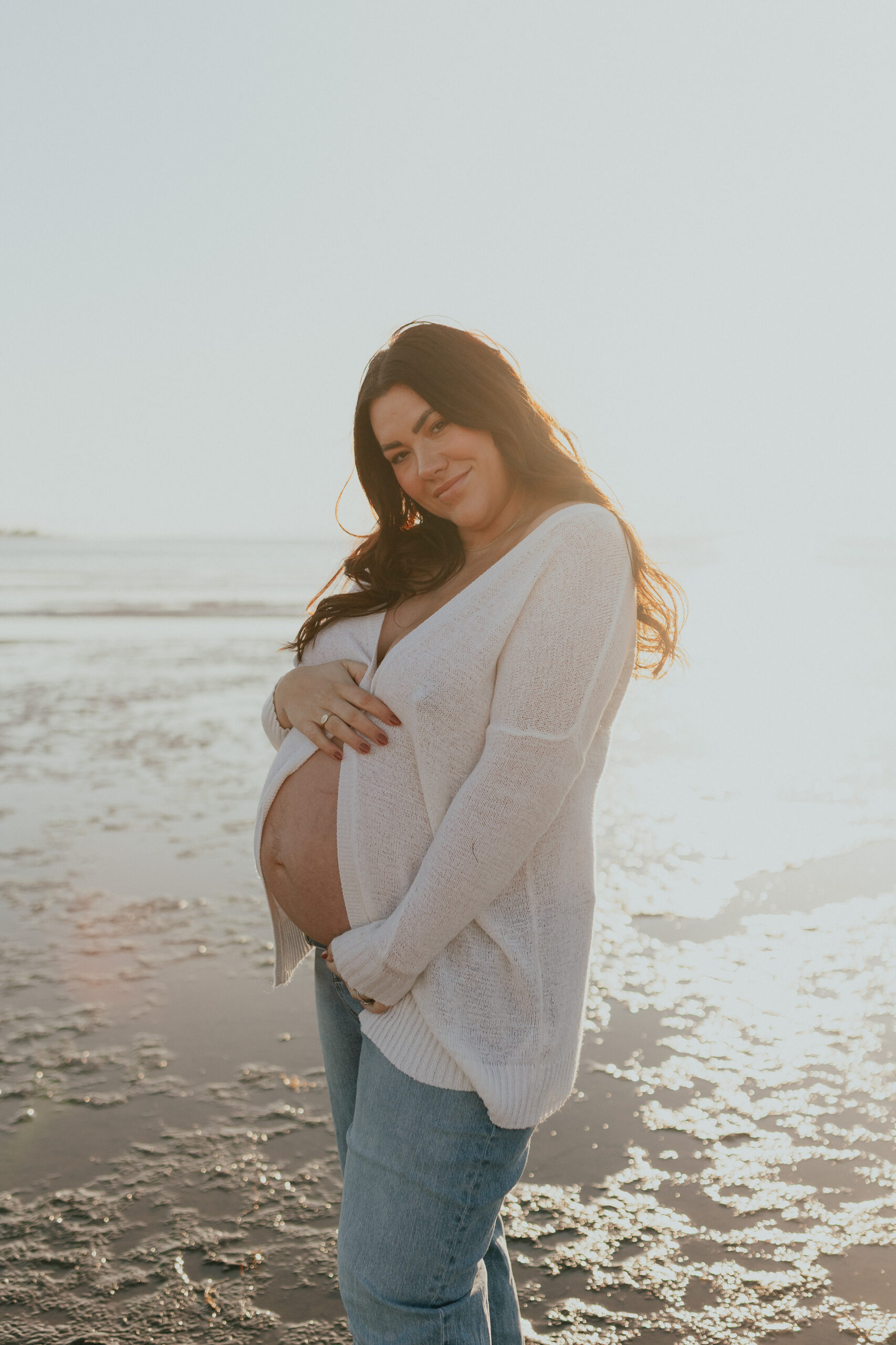 Photo of a pregnant woman smiling softly at the camera while holding her baby bump with the sun setting behind her