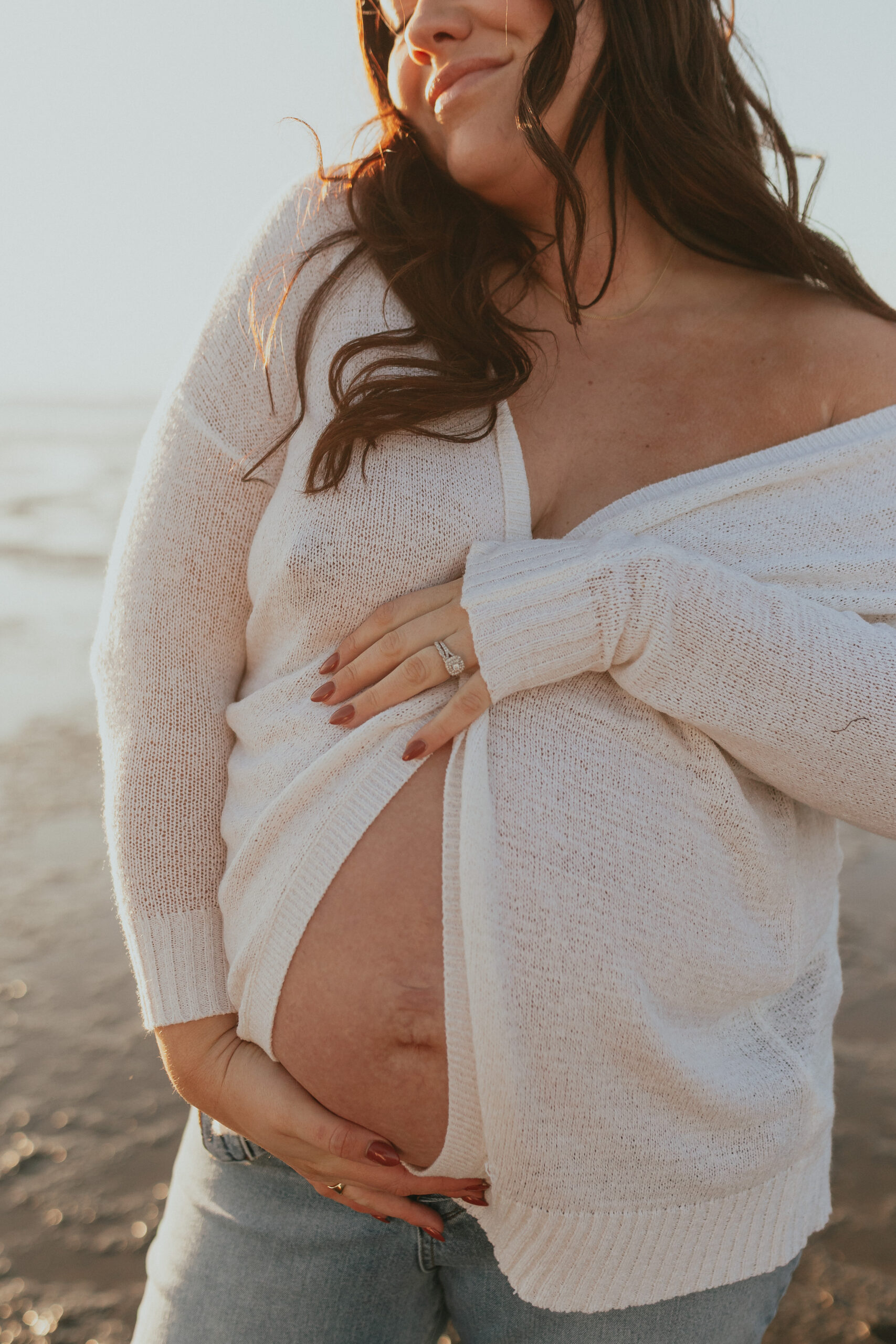 photo of a pregnant woman holding her baby bump while standing on the beach. You can barley see her face but her eyes are closed and she is smiling. Taken by a Vancouver Maternity Photographer.