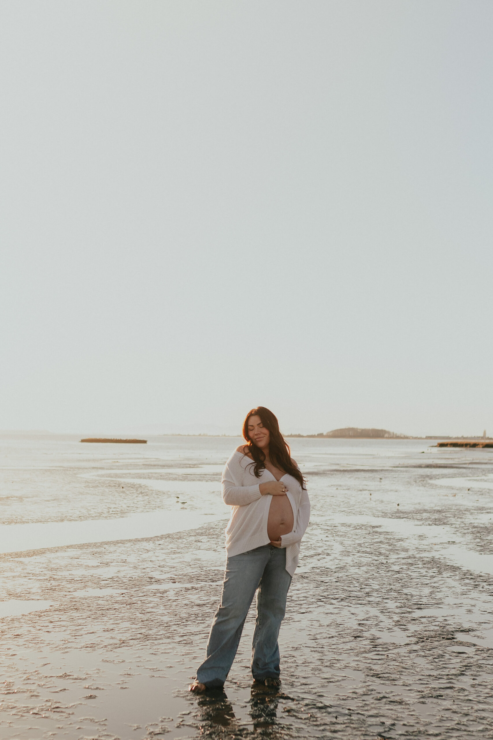 Photo of a pregnant woman standing on the beach with the sun setting behind her while she softly smiles at the camera and holds her baby bump