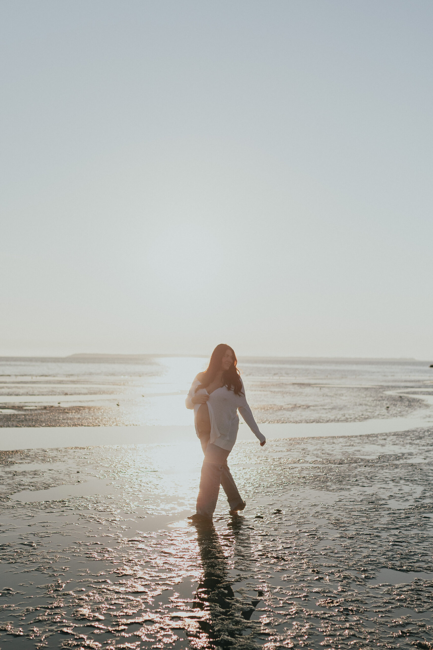 Photo of a pregnant woman on the beach with the sun setting behind her while she walks on the wet sand. taken by a vancouver maternity photographer