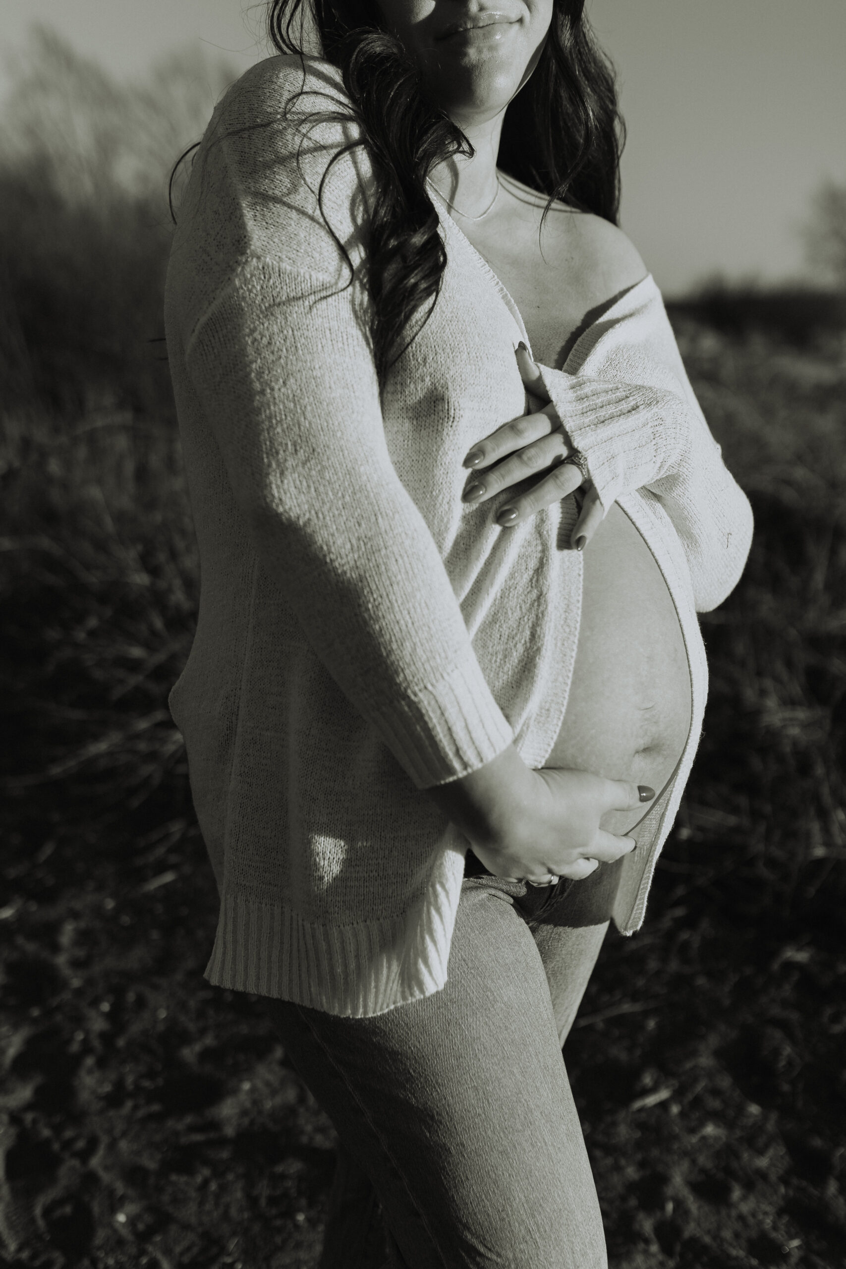 black and white photo of a pregnant woman holding her baby bump with the focus of the photo being on her belly, but you can see her soft smile at the top of the photo