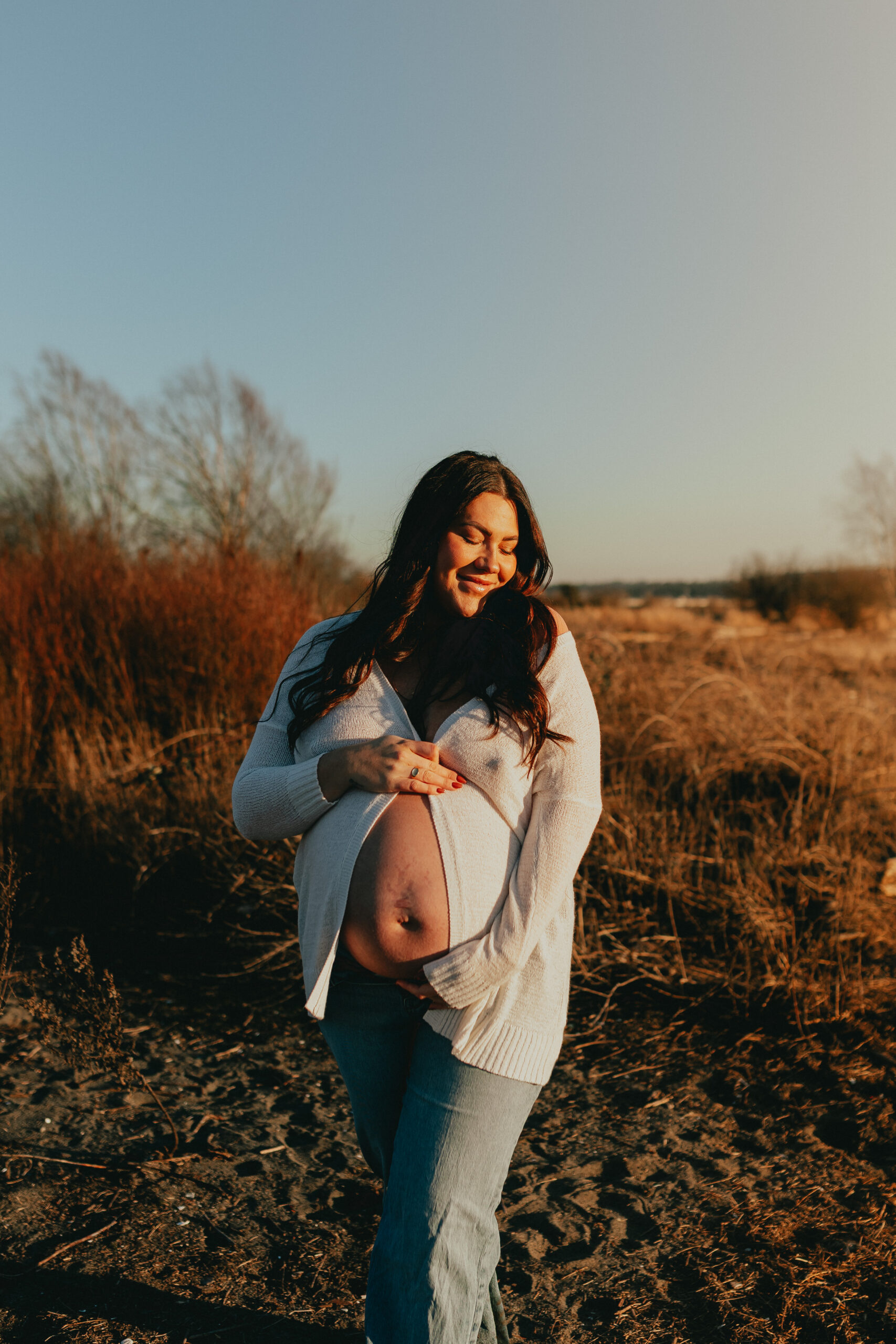 Photo of a pregnant woman with the sun on her face and holding her baby bump while looking to the side and smiling