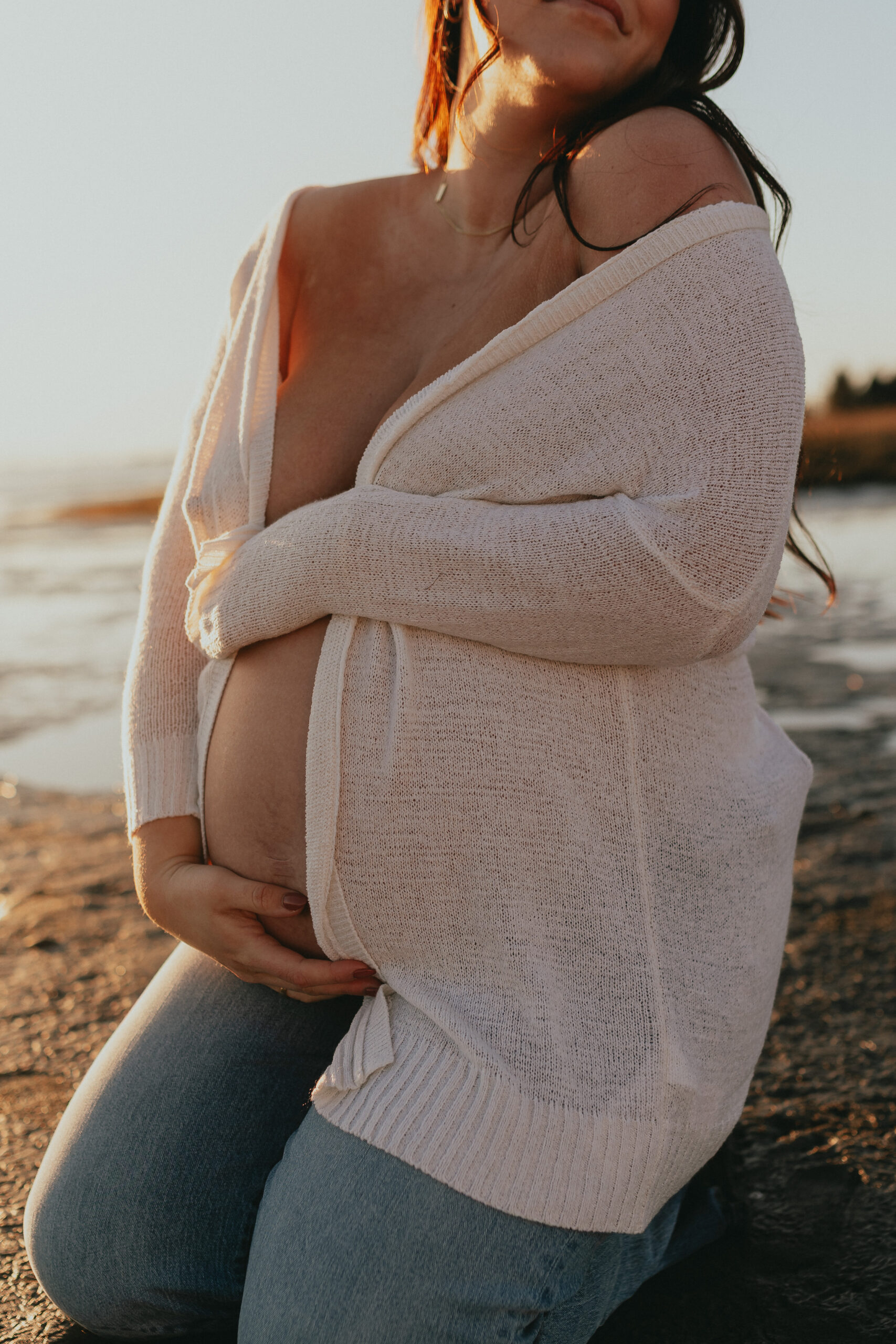 Close up photo of a pregnant woman kneeling on the sane on the beach holding her bare baby bump