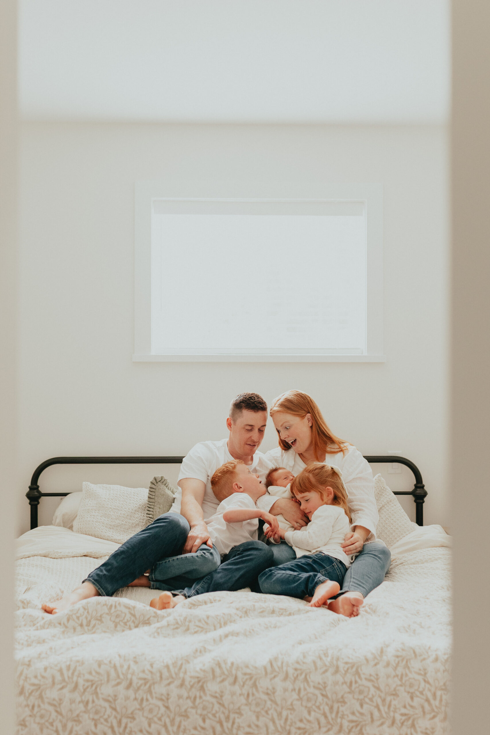 Photo of a family of 5 sitting on the big bed while everyone is looking at each other smiling and mom is holding the newborn baby
