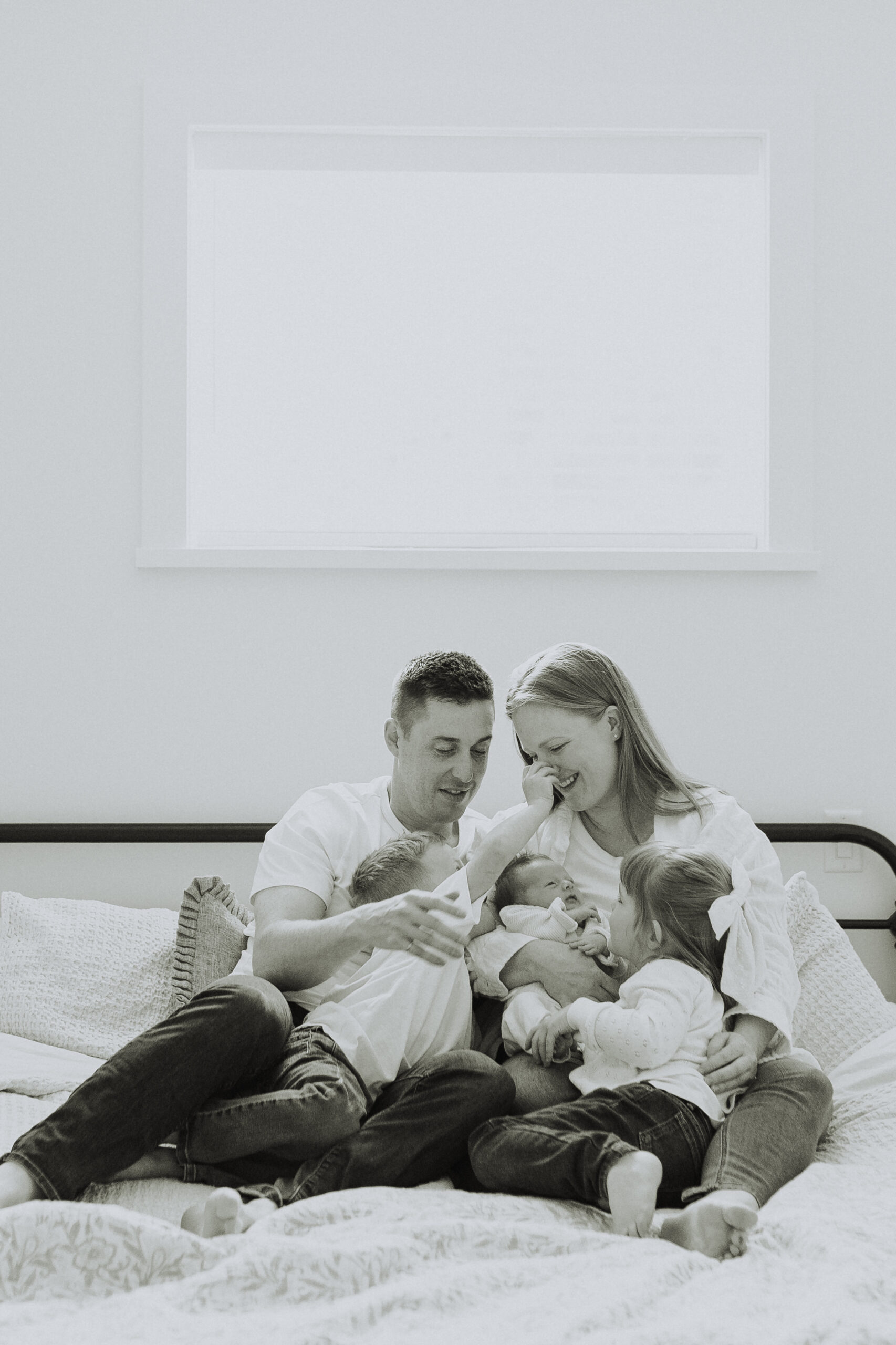 Black and white photo of a family sitting on the big bed looking at the newborn baby taken for newborn family photos