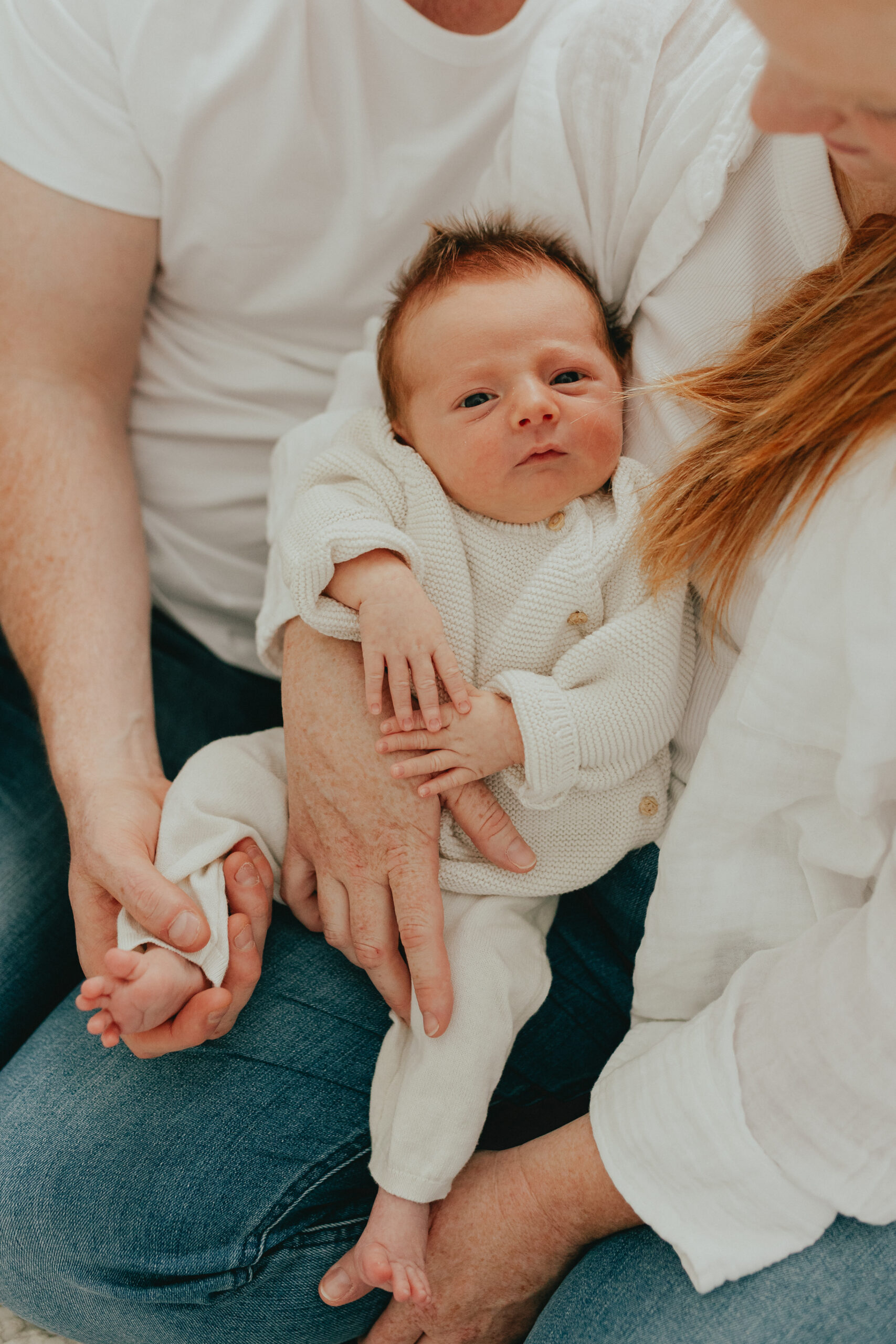 Photo of a newborn baby being cradled in between mom and dad sitting on their bed taken for newborn family photos
