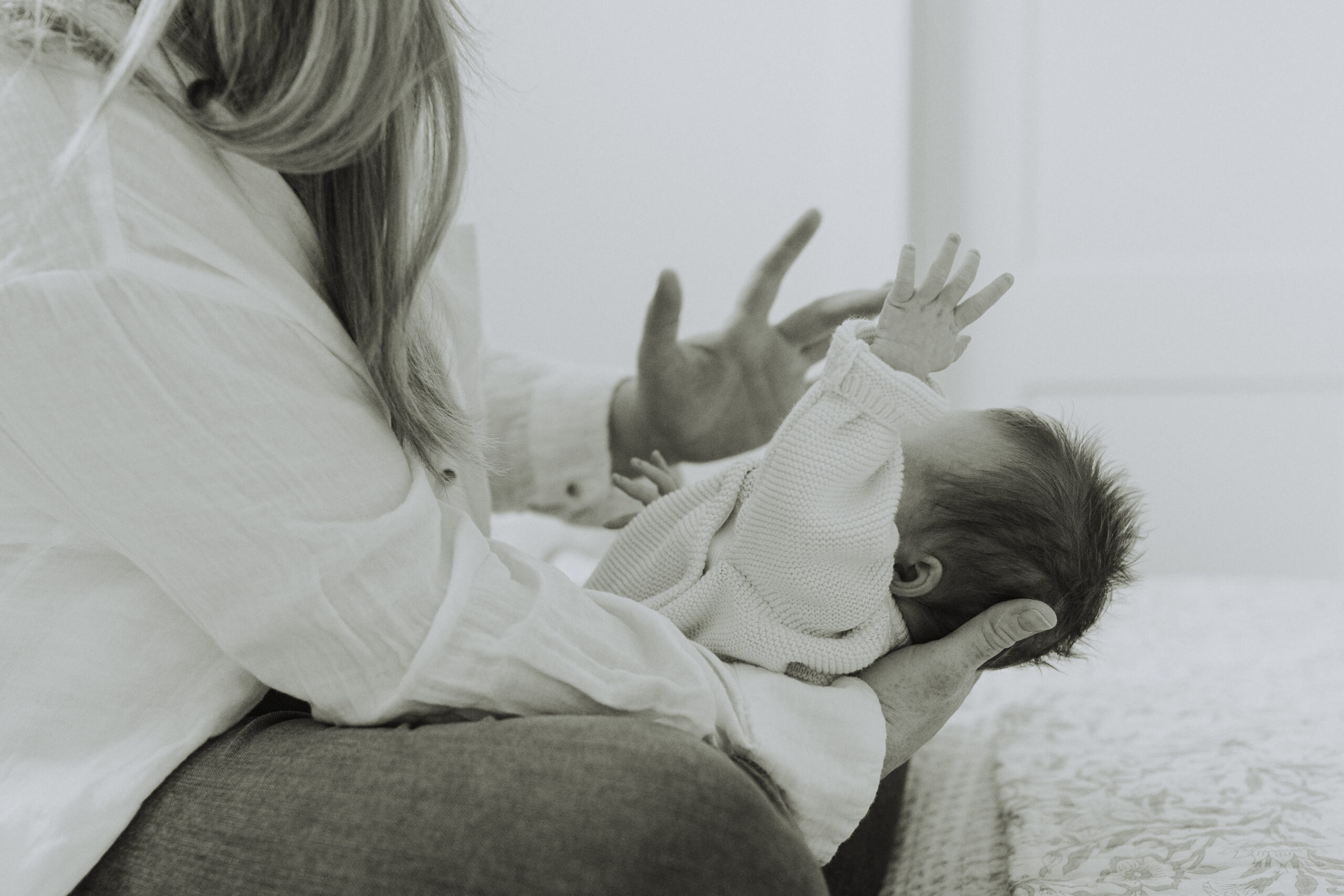 Black and white photo of a mom holding her newborn baby. The baby has their hand in the air and mom is stroking the side of his face. Taken for newborn family photos