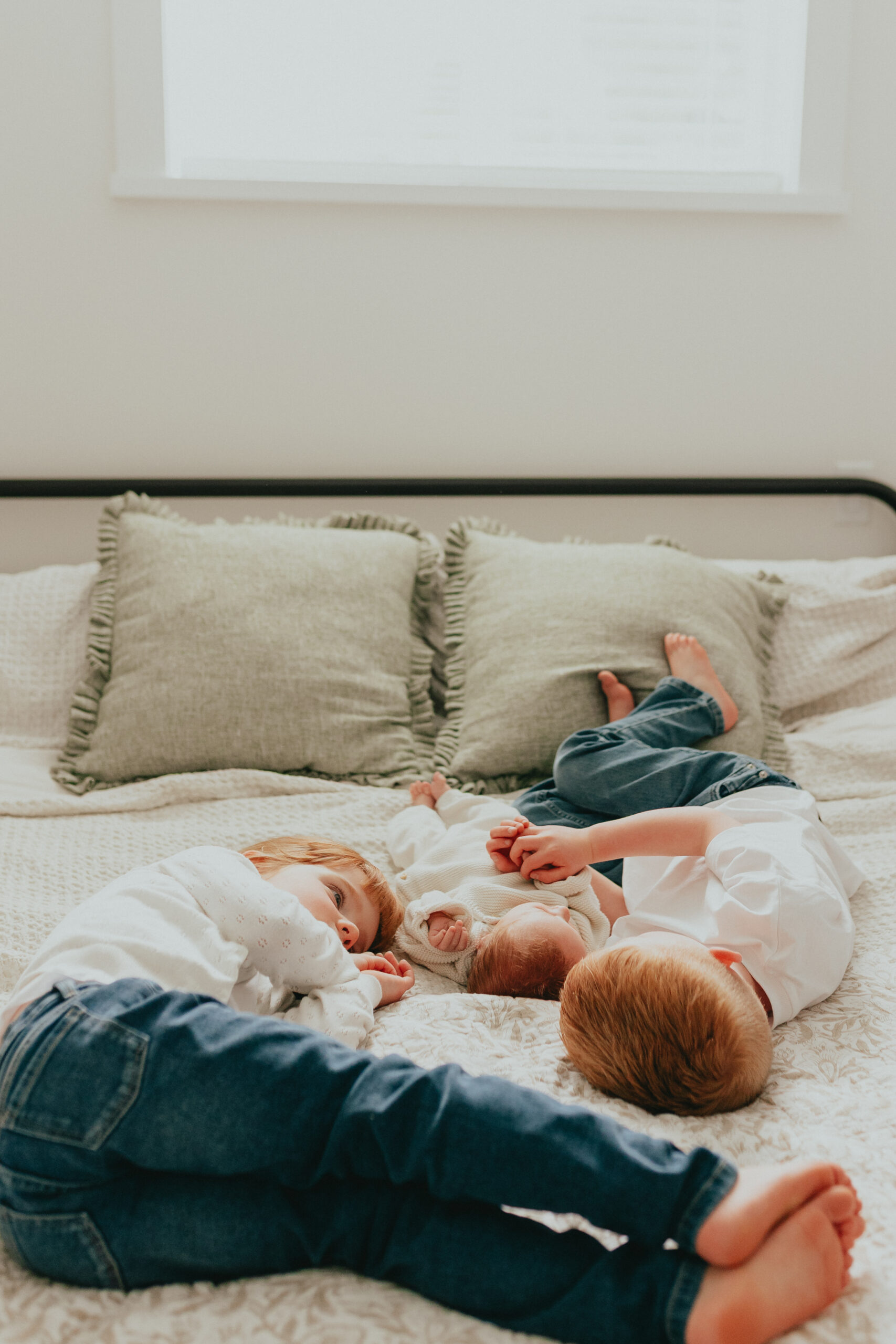 Photo of a big brother and sister laying on the big bed with their newborn baby brother laying in between them and they are looking at him