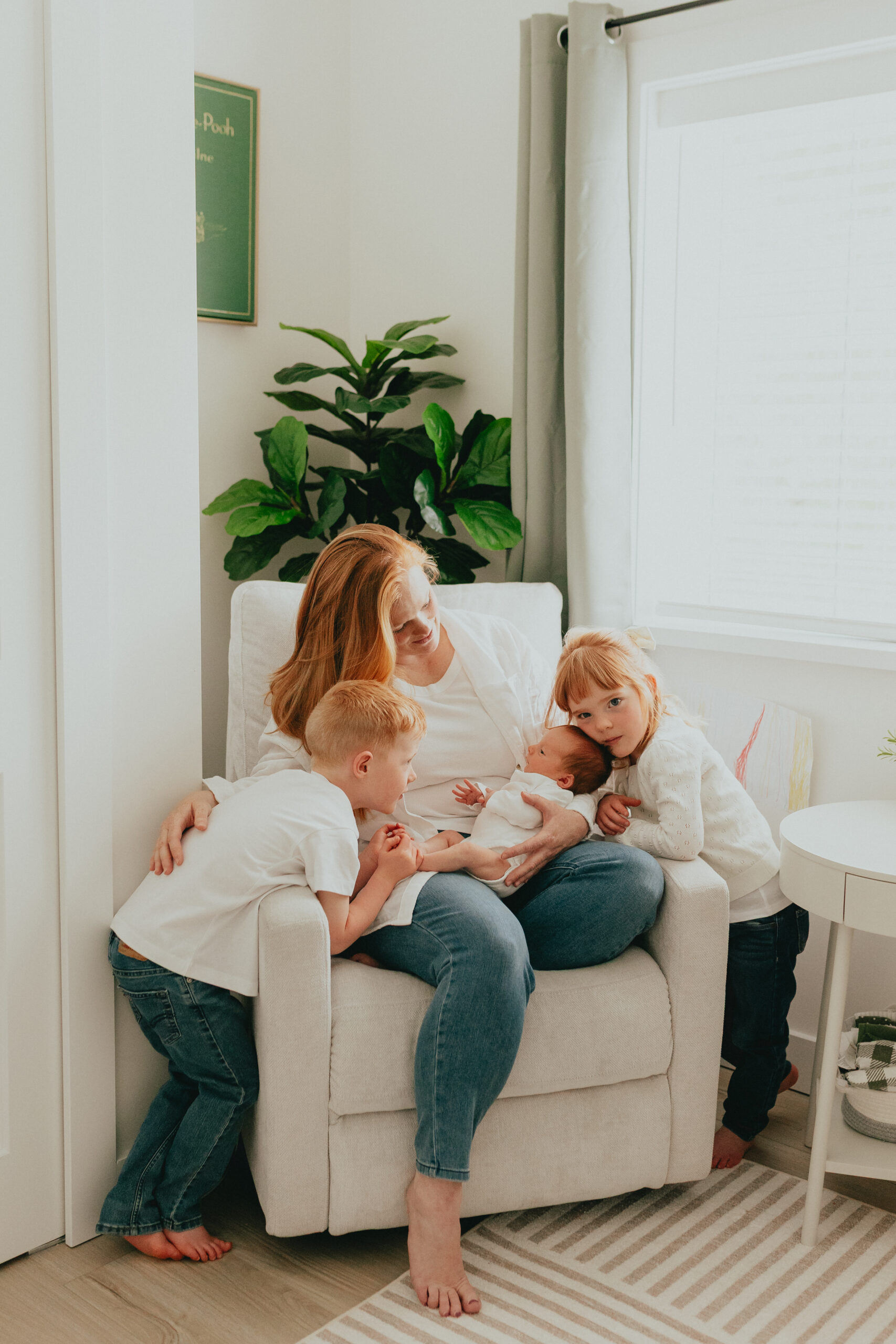 Photo of mom sitting on the nursery chair holding a newborn baby while big sister and big brother lean over looking at the baby
