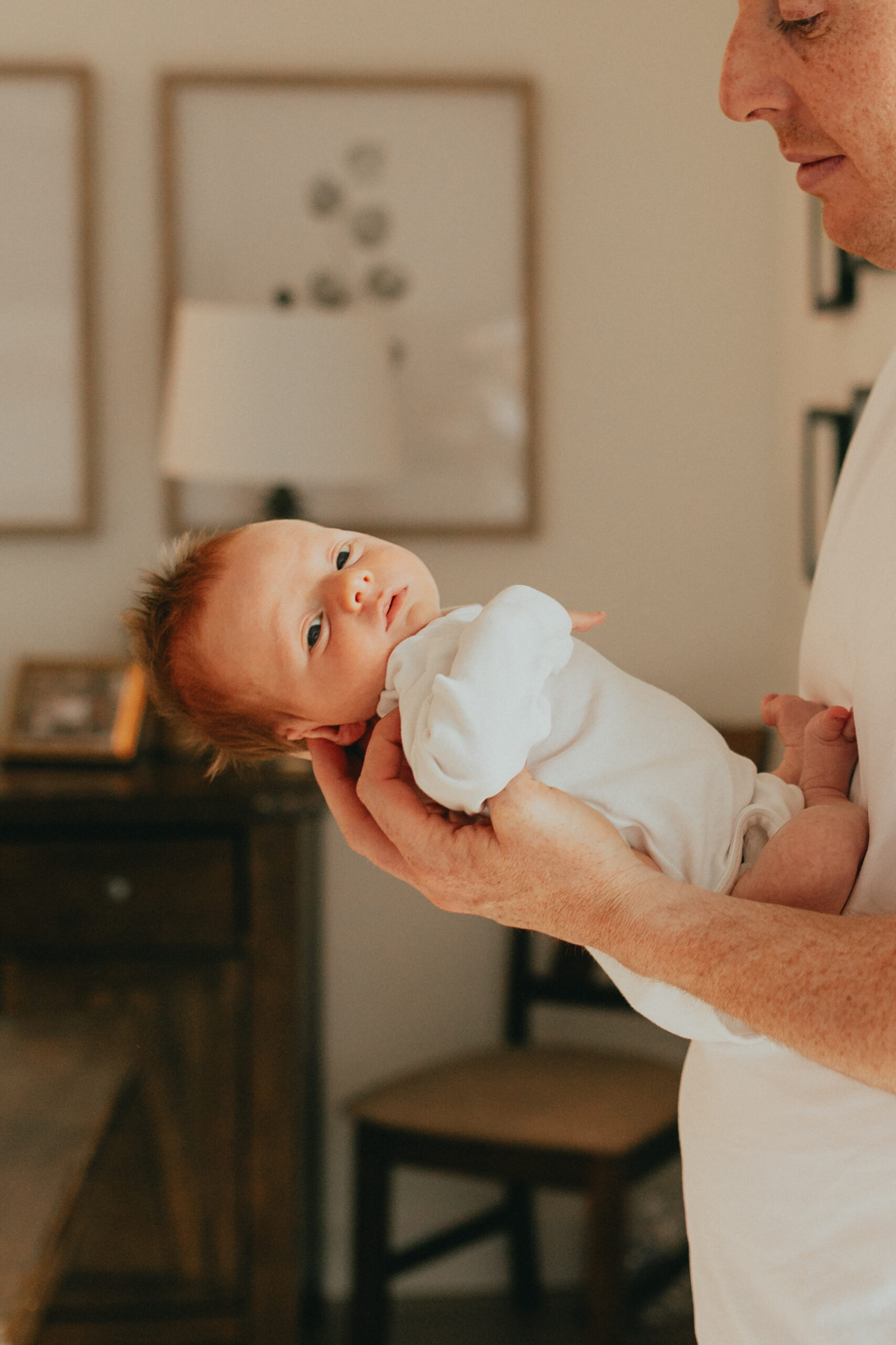 Photo of a dad holding the newborn baby and the baby is turning and looking at the camera