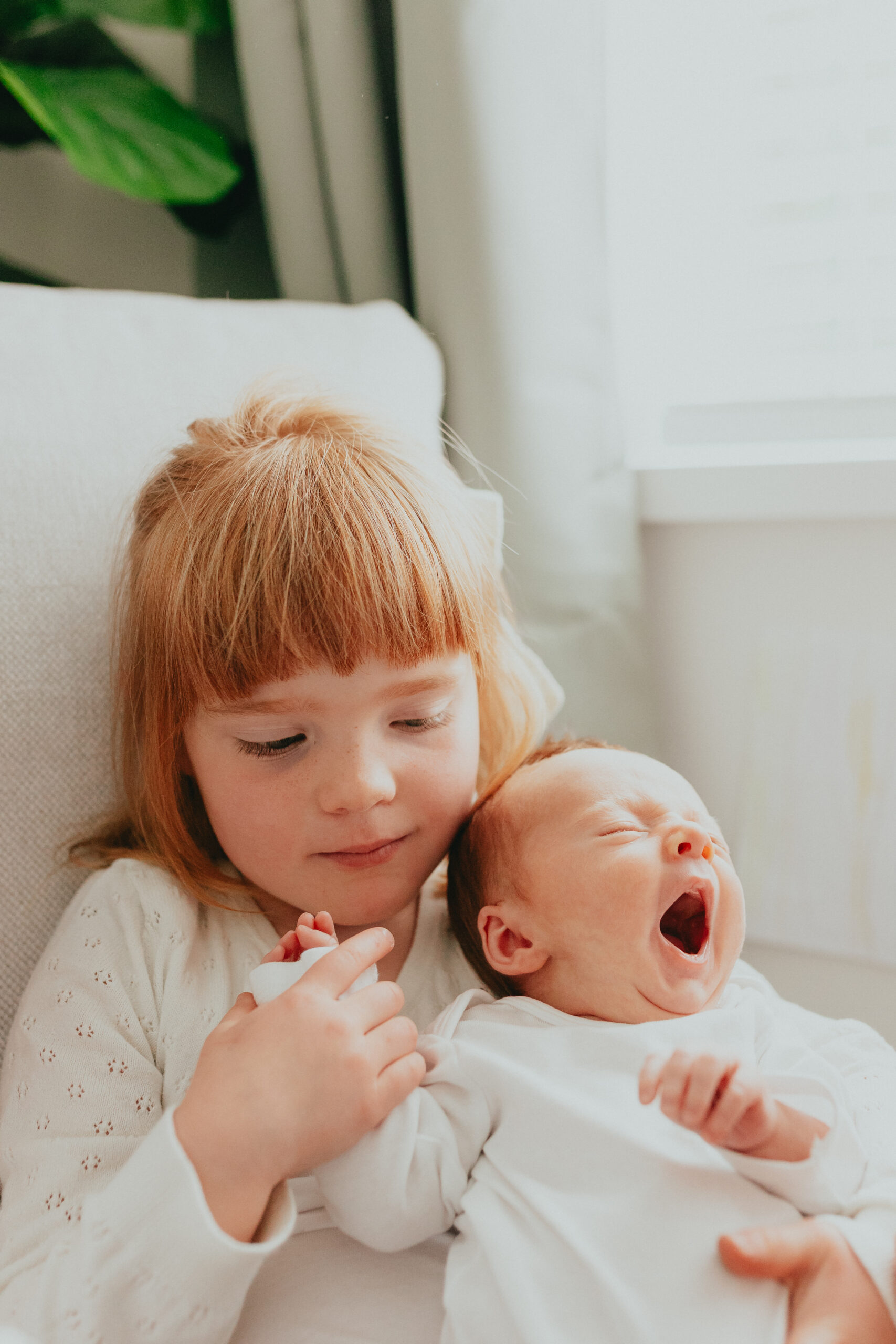 Photo of a big sister holding her baby brother while she looks down at him yawning taken for newborn family photos