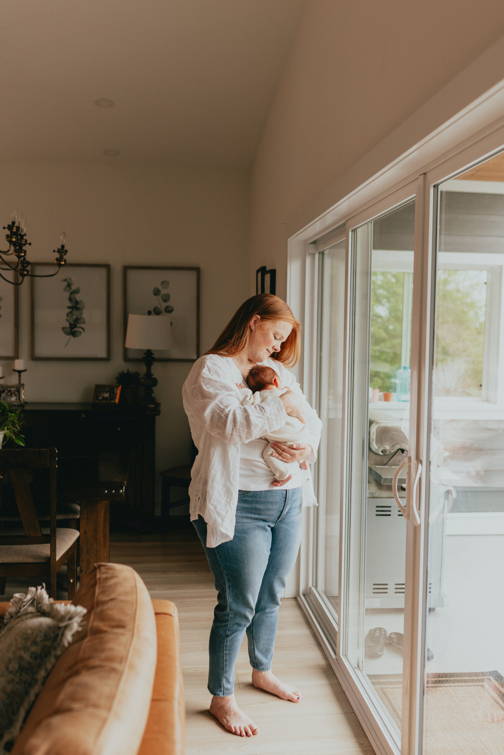 Photo of a mom standing by the sliding glass door holding and looking at her newborn baby