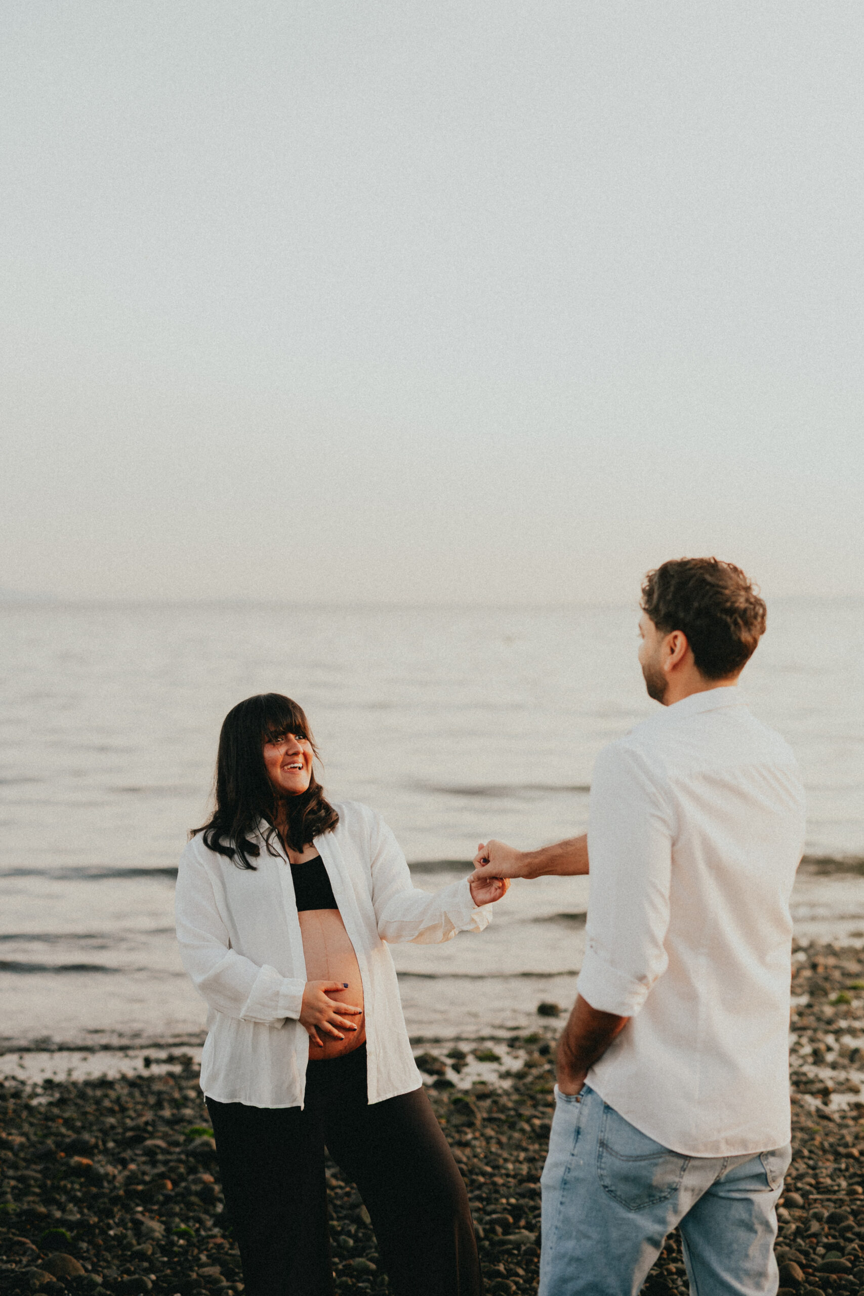 Photo of a husband and wife on their vancouver babymoon on the beach with the ocean behind them. the wife is looking at the husband smiling