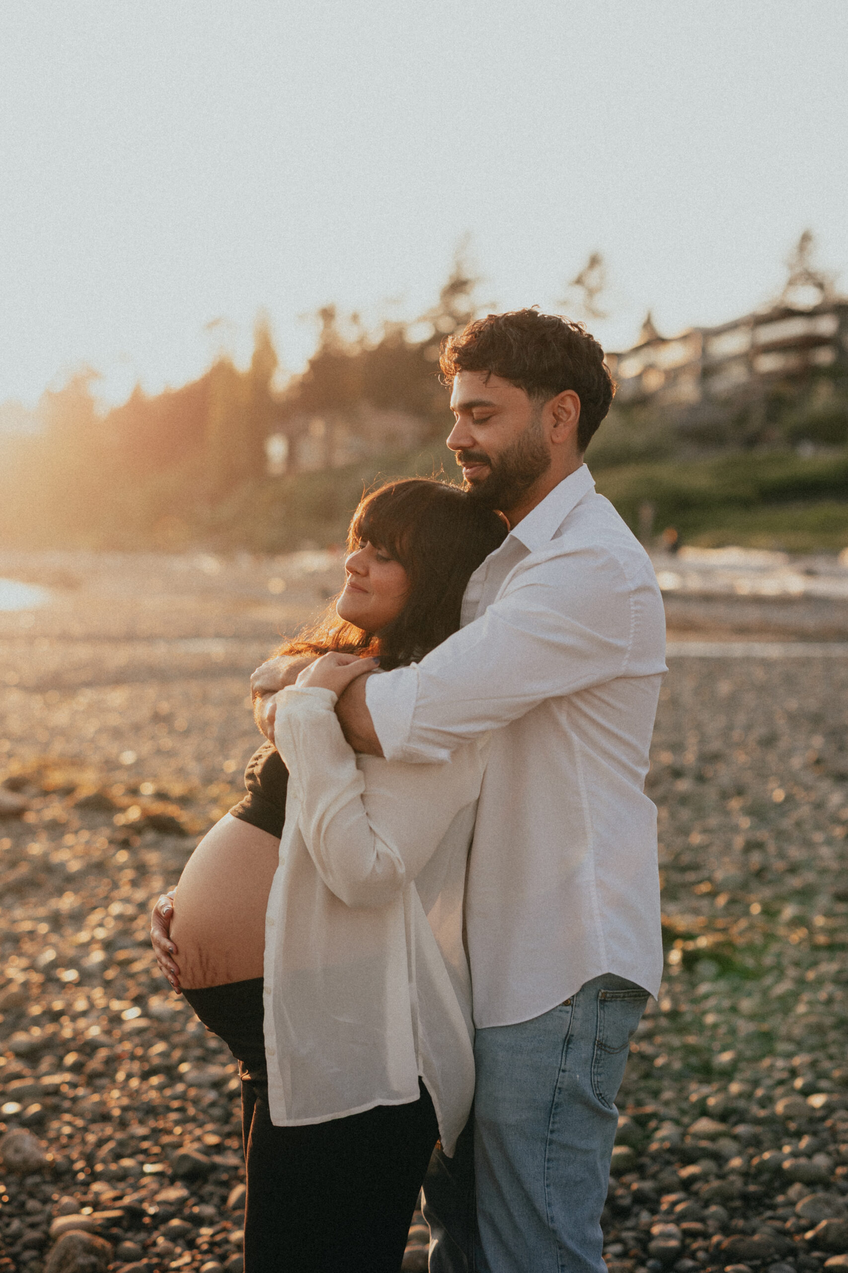 A couple on their Vancouver Babymoon on the beach. The husband is hugging his pregnant white from behind and they both have their eyes closed