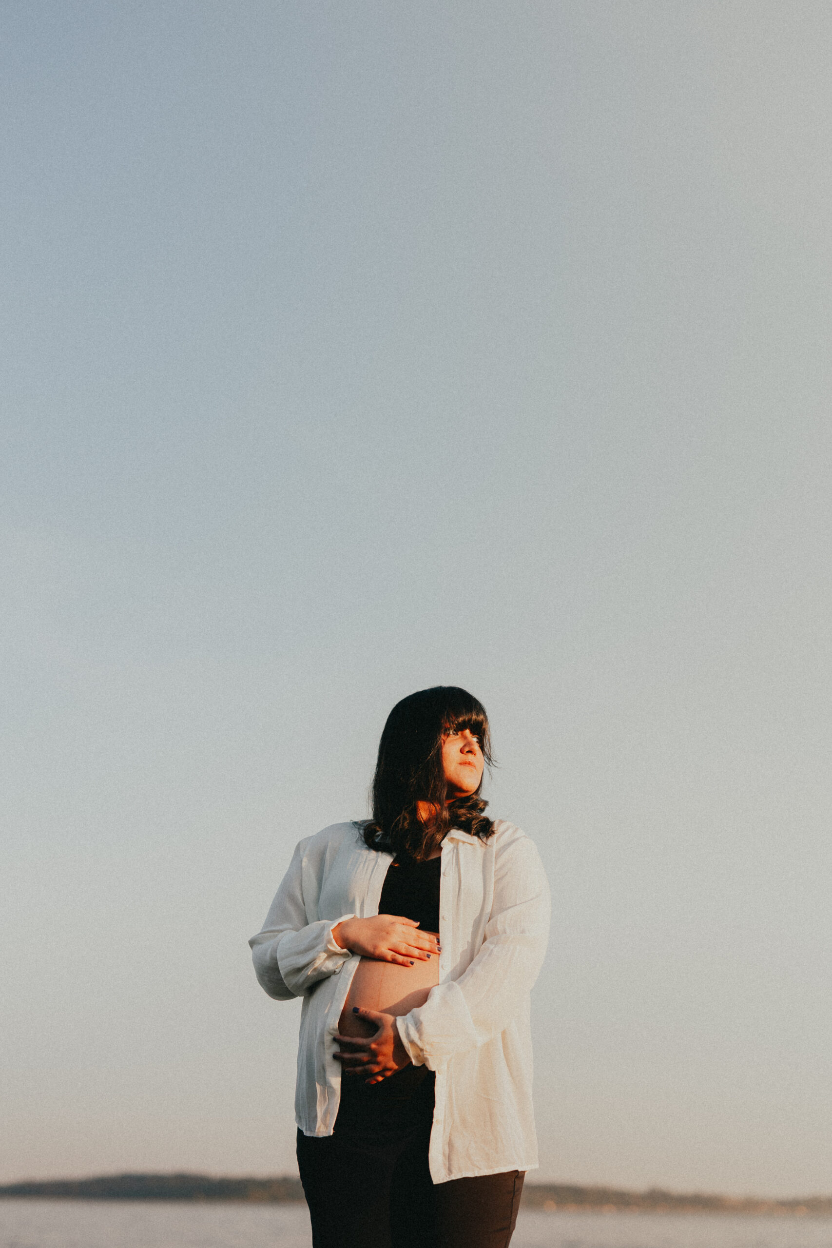 Photo of a pregnant woman on a Vancouver Babymoon standing on the beach holding her baby bump and looking off into the distance