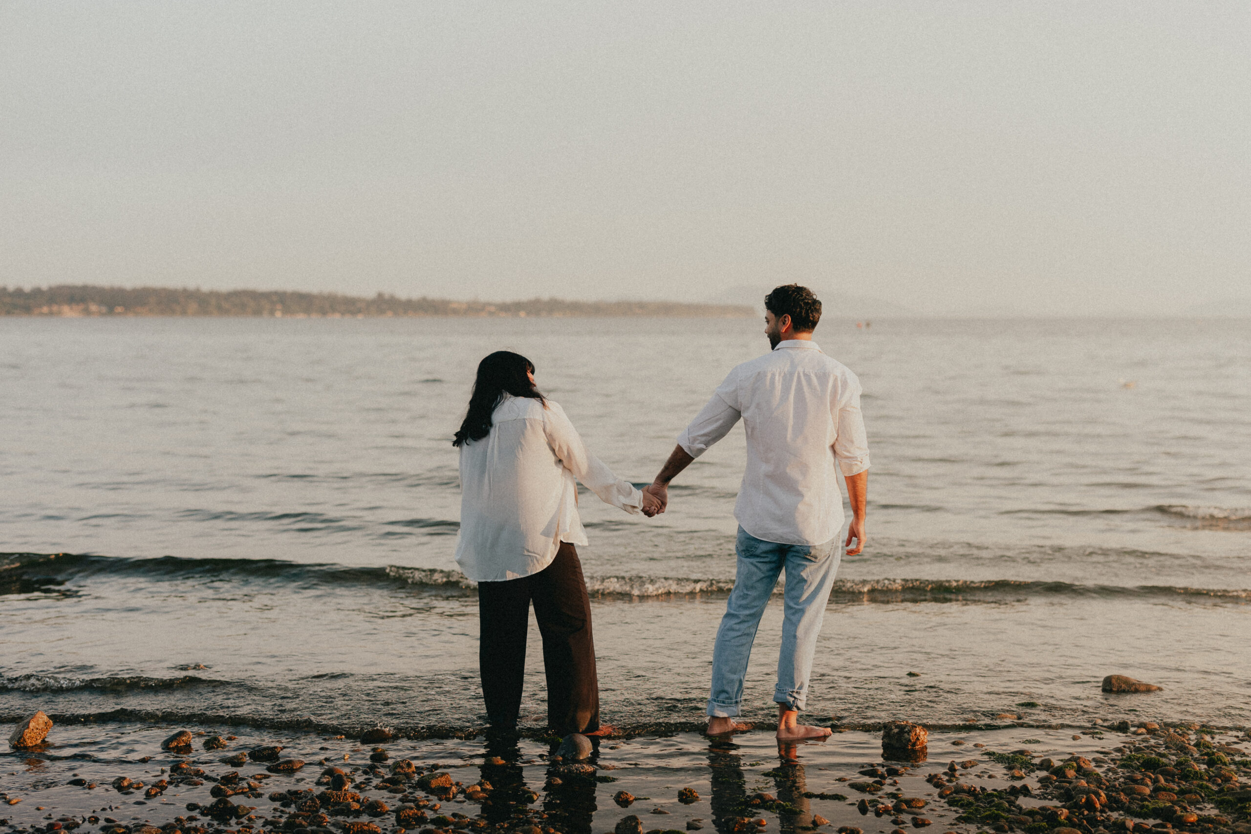 Photo of a husband and wife standing in the ocean holding hands while looking at each other with their backs to the camera