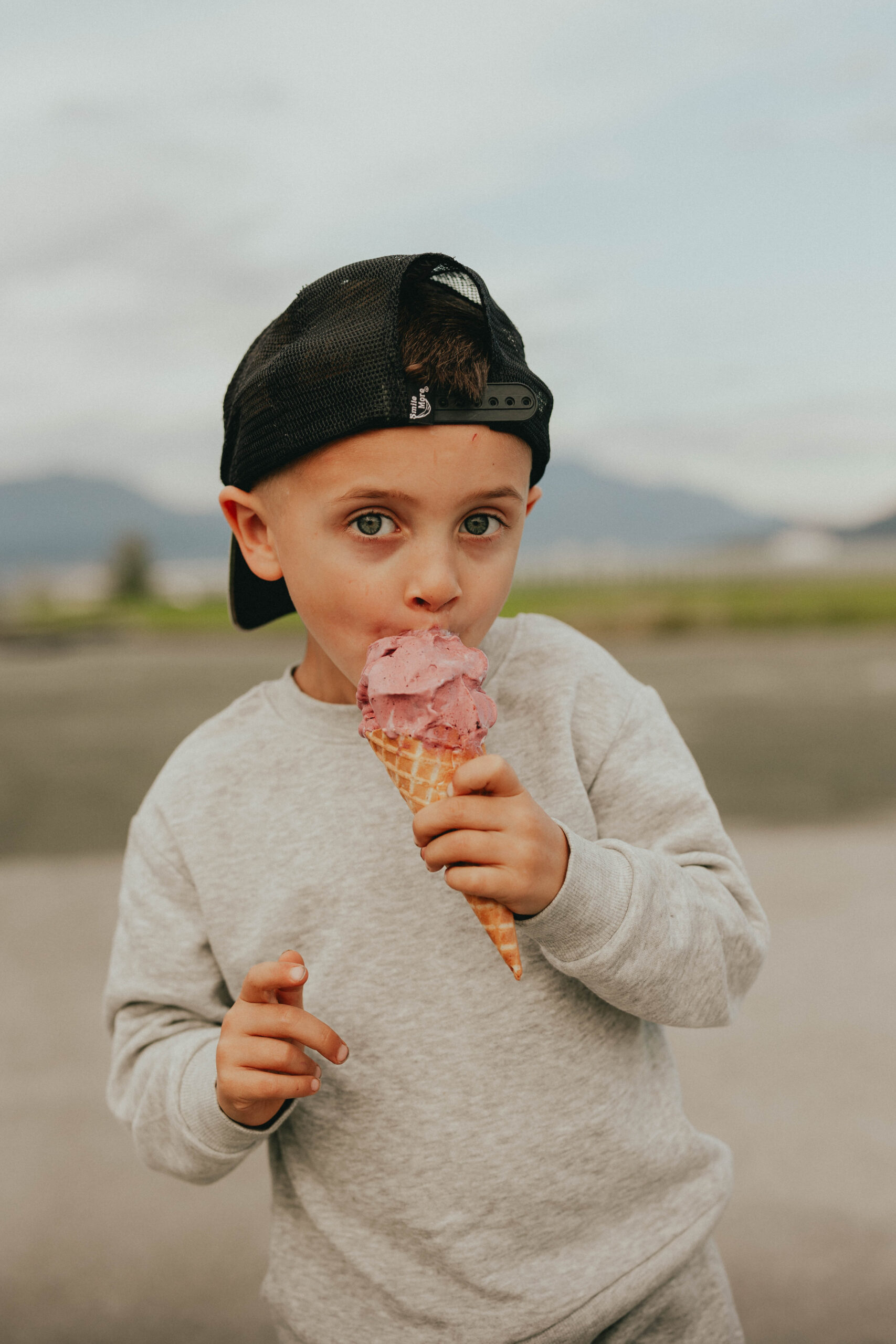 Photo of a boy wearing a backwards hat licking his ice cream while looking at the camera