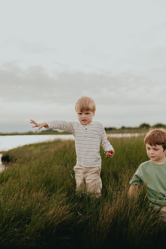 Photo of 2 young boys playing by the ocean, one sitting in the grass and one standing throwing rocks in the ocean
