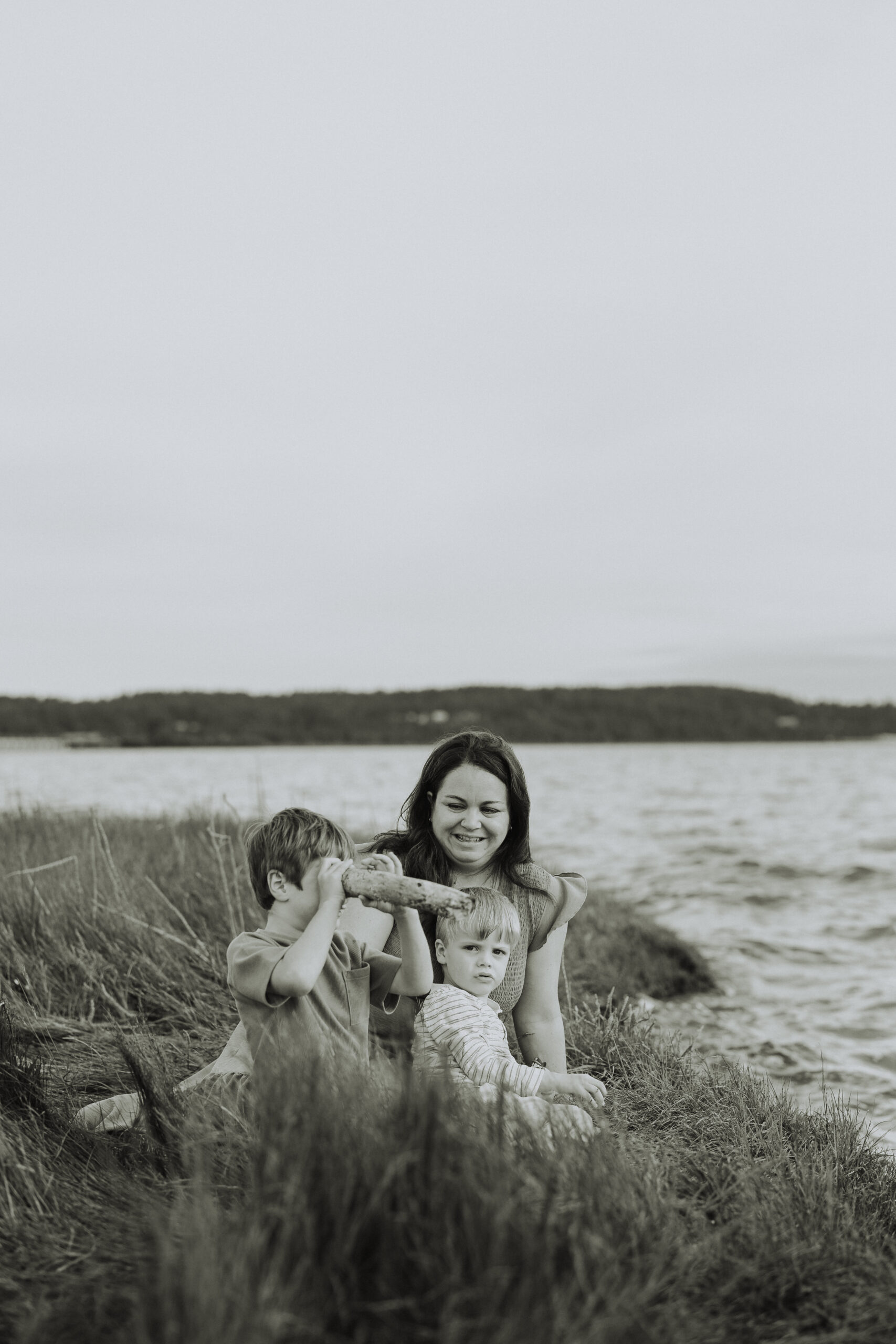 Black and white photo of a mom and her 2 young sons sitting by the ocean playing with rocks and sticks. Used in a blog about langley pools