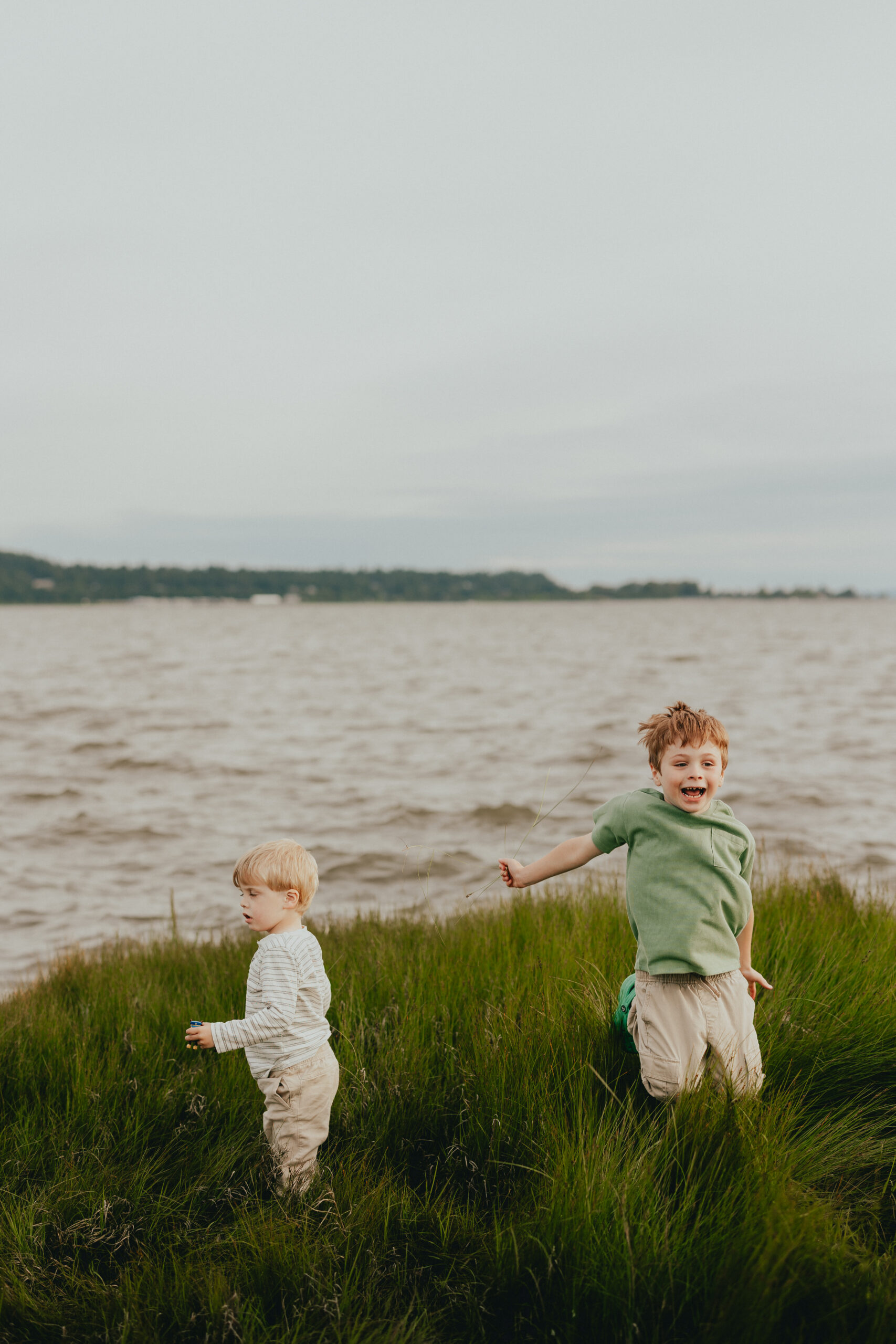 Photo of 2 young boys playing by the ocean, one facing the water throwing in rocks and one facing the camera jumping up in the air