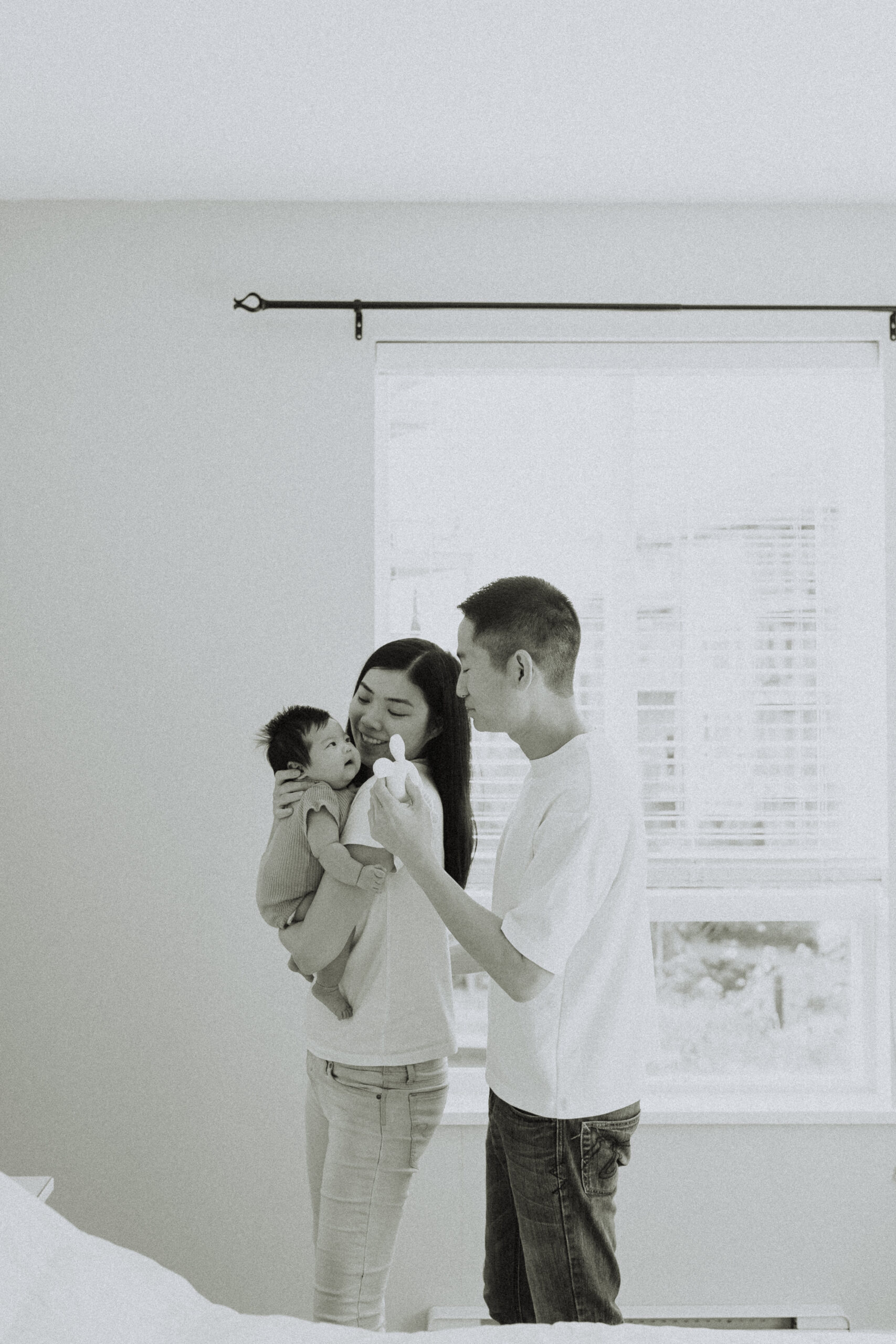 Black and white photo of a mom holding her baby girl over her shoulder and dad holding a toy for her