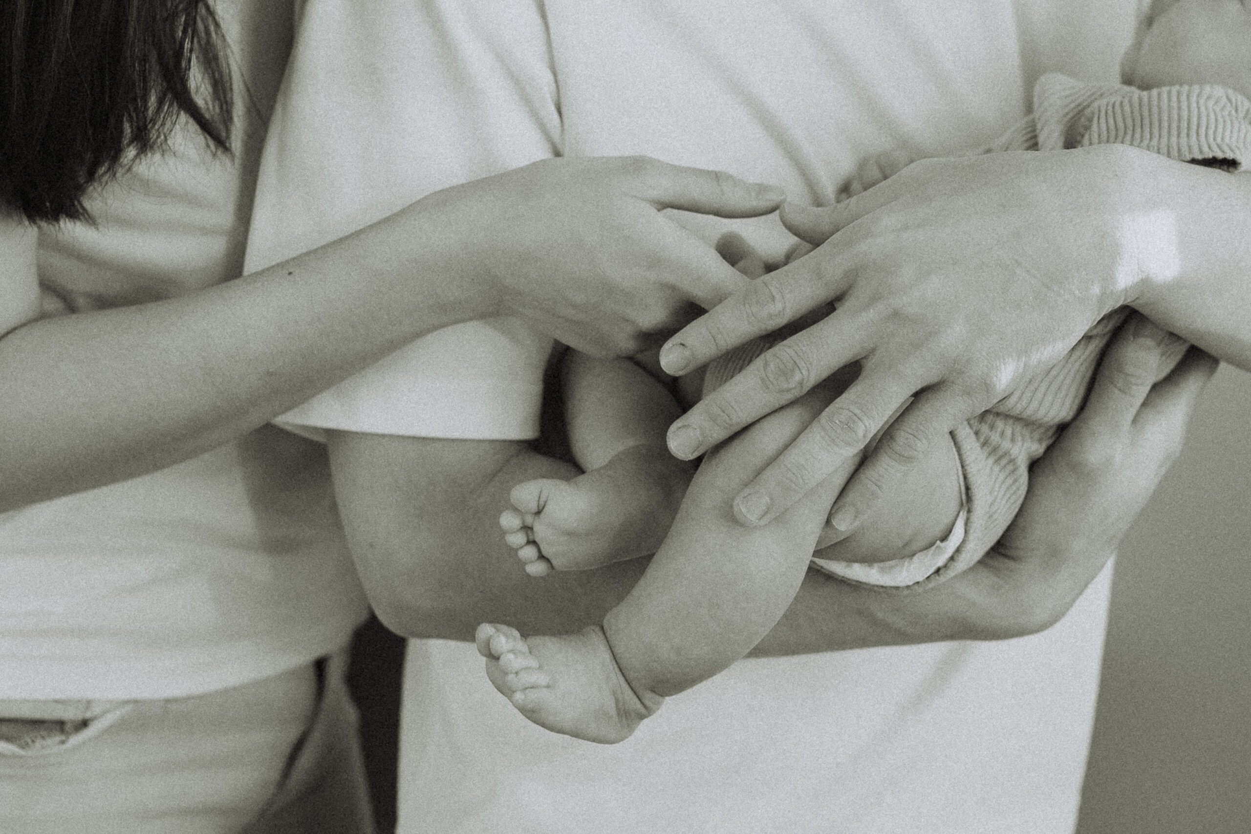 Black and white photo of a dad holding his newborn baby close up on his arms and the baby's legs and mom holding the baby's hand