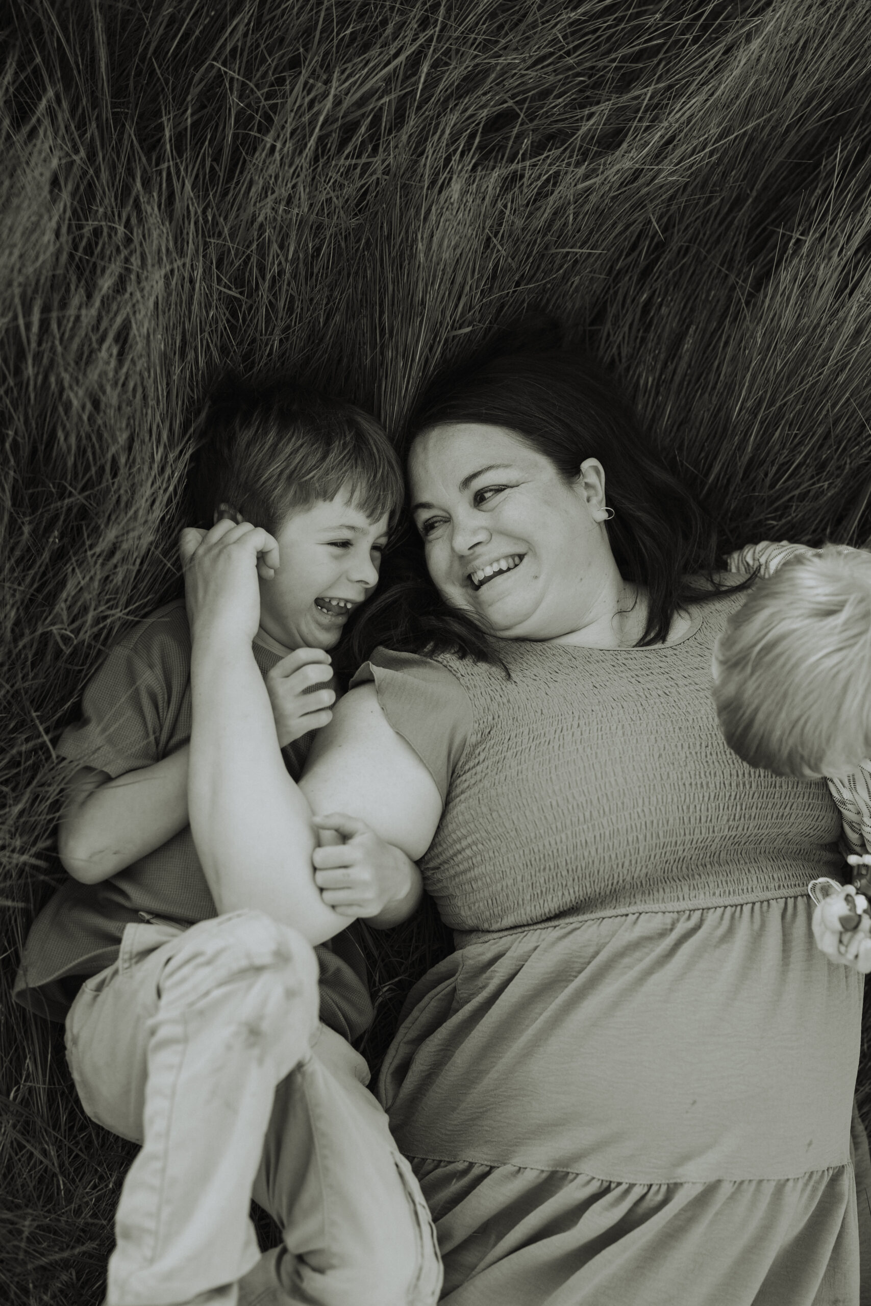 Black and white photo of a mom laying in the grass with her young sons playing and smiling at each other