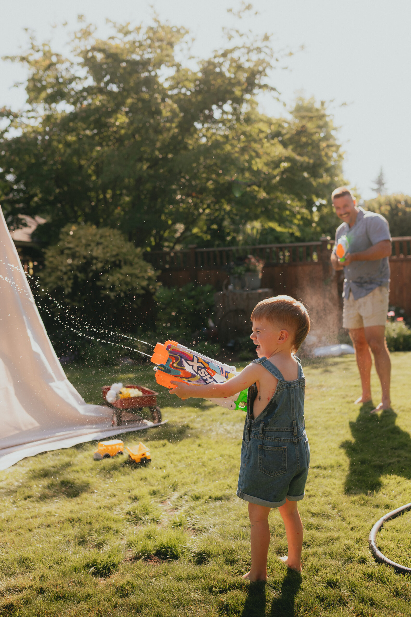 Photo of a boy with a water fun squirting it at someone off camera while his dad in the distance squirts him with a water gun