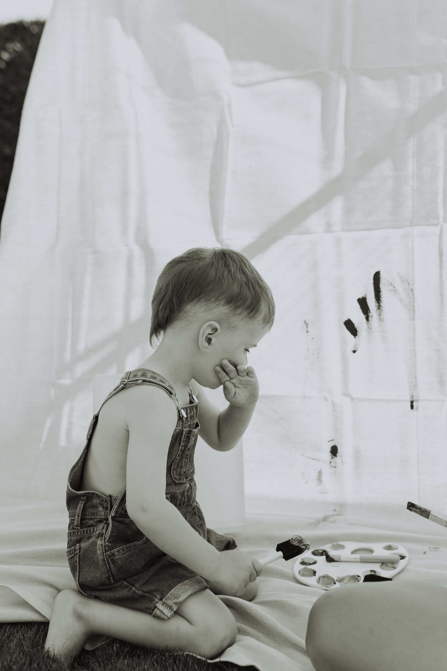 black and white photo of a boy sitting in front of a big canvas wiping paint off his cheek taken by a documentary photographer