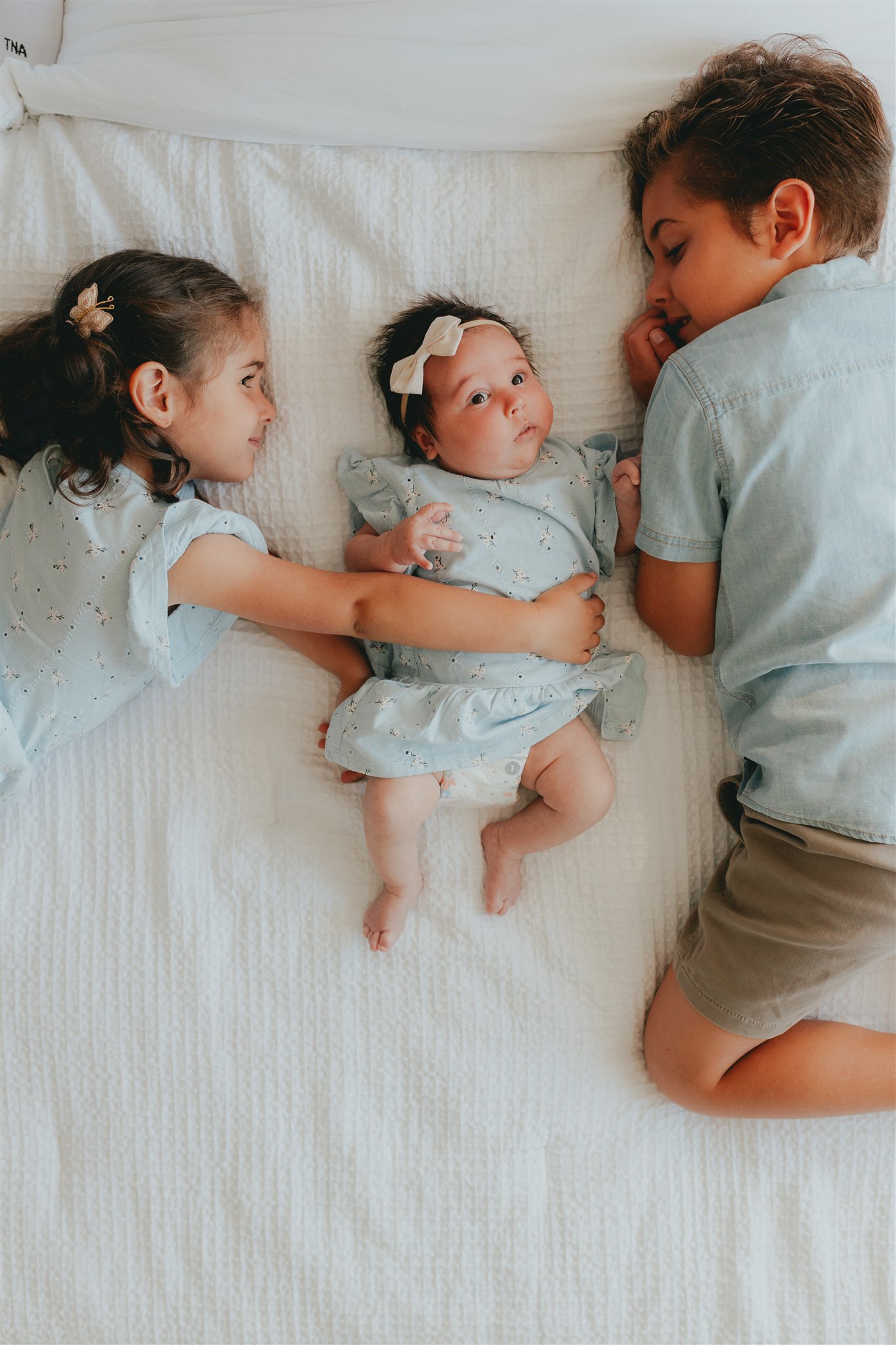 An older brother and sister lay on the bed with their baby sister in the middle. They are looking at her with the photo taken from above. For a blog about newborn photography with siblings.