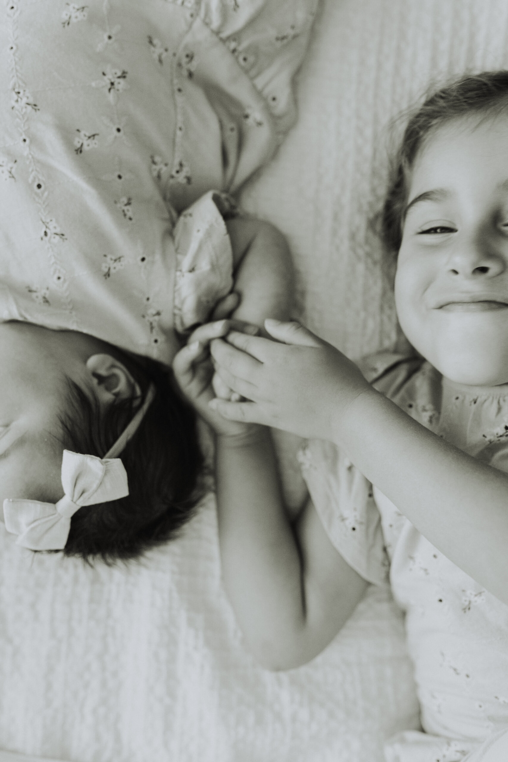 Black and white photo of a little girl laying on the bed beside her baby sister holding her hand smiling up at the camera