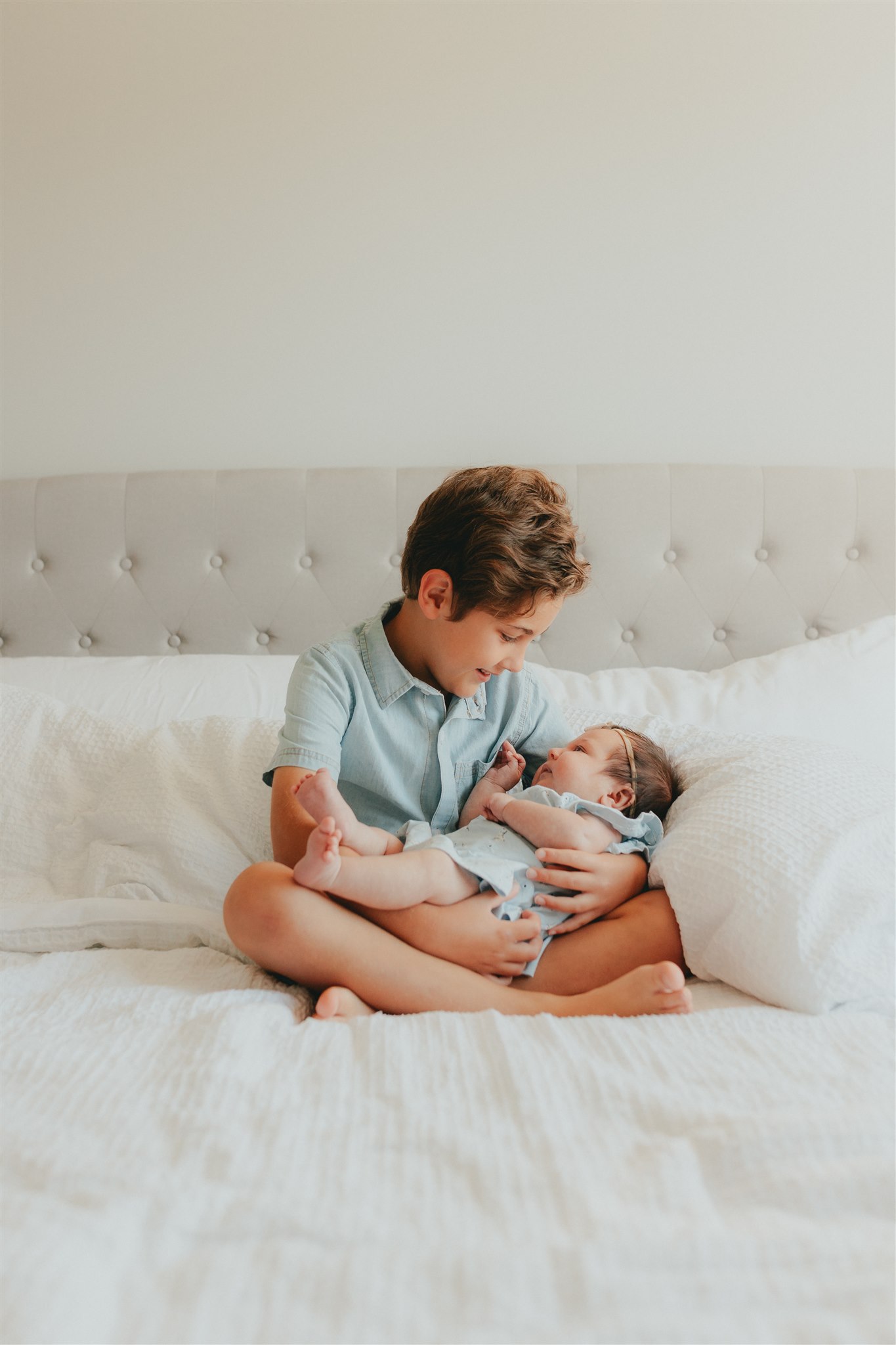 Photo of a boy sitting on the bed holding his baby sister and looking down at her. For a blog about newborn photography with siblings